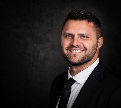 Smiling man in a black suit and tie seated in a dimly lit office with warm accent lighting