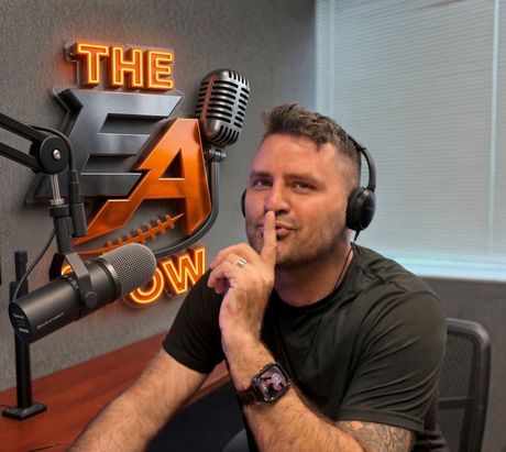 Podcast host at a desk with microphone and headphones, making a hush gesture beside a neon studio sign.