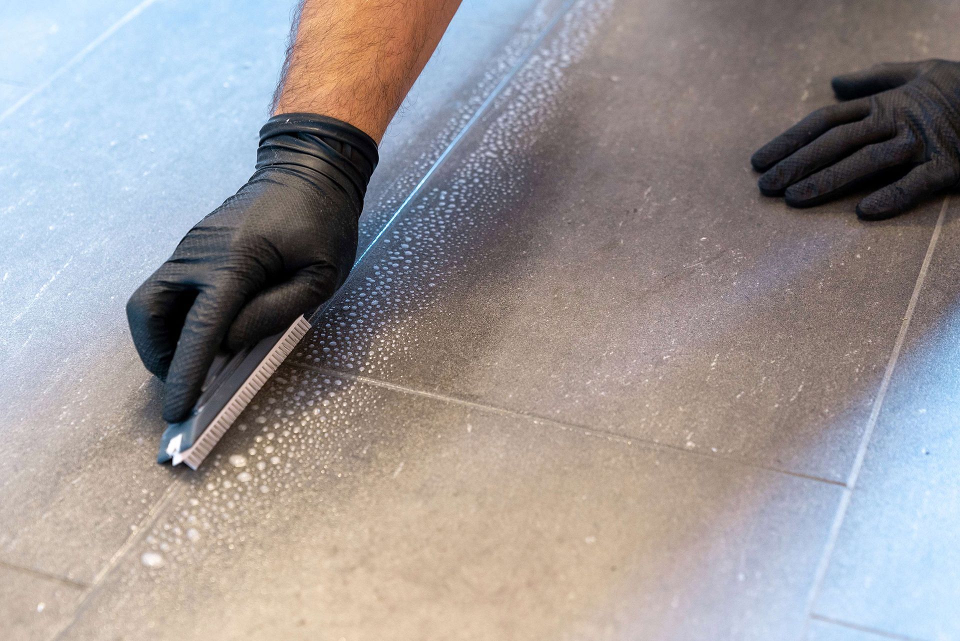 A person wearing black gloves is cleaning a tile floor with a sponge. A person wearing black gloves is cleaning a tile floor with a sponge.
