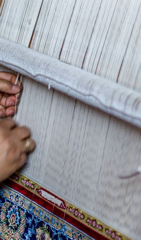 Hands weaving white threads into a colorful rug on a loom.