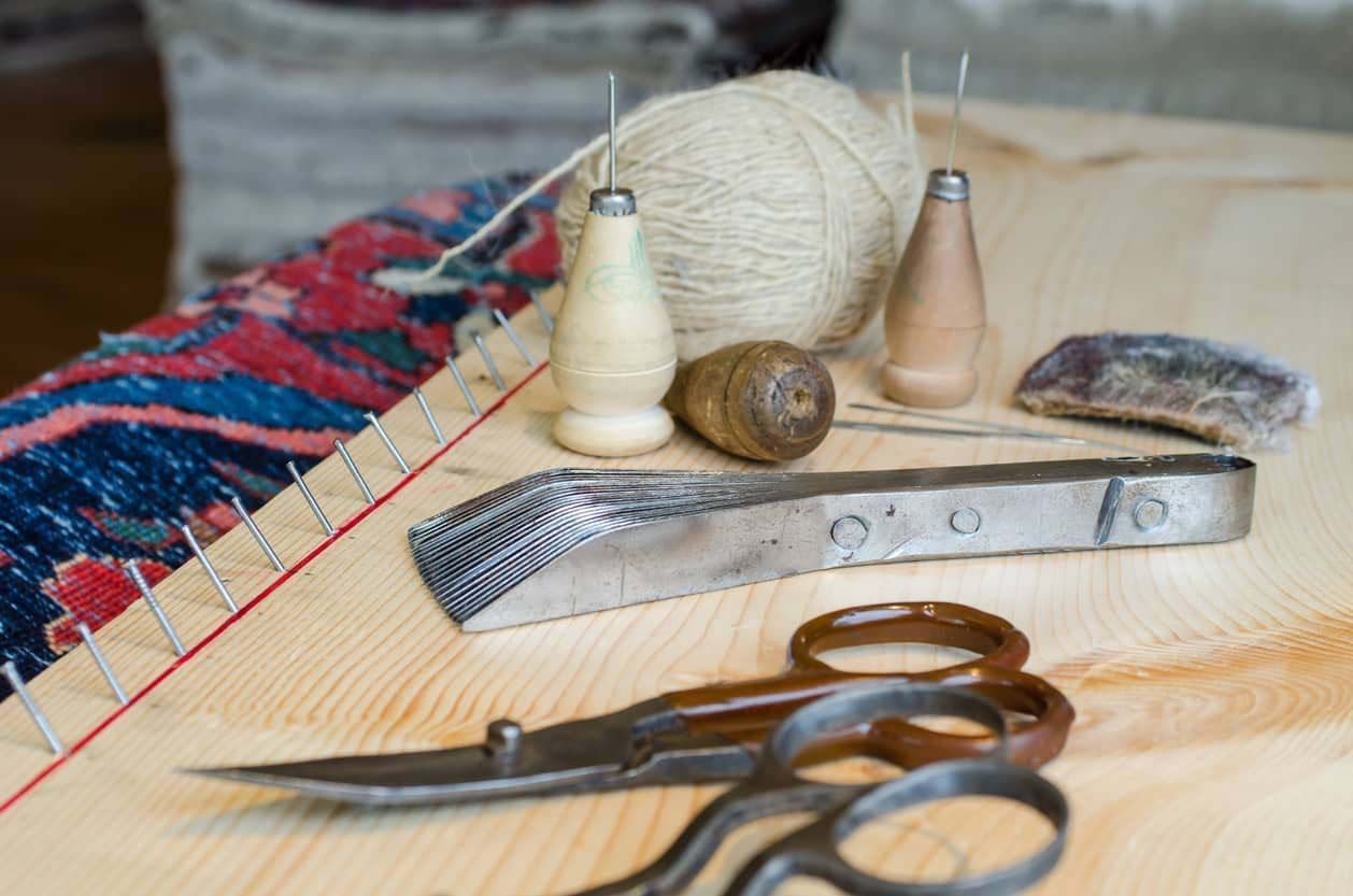 Tools for rug making: scissors, thread, wooden tools, and a metal clamp on a wooden frame.