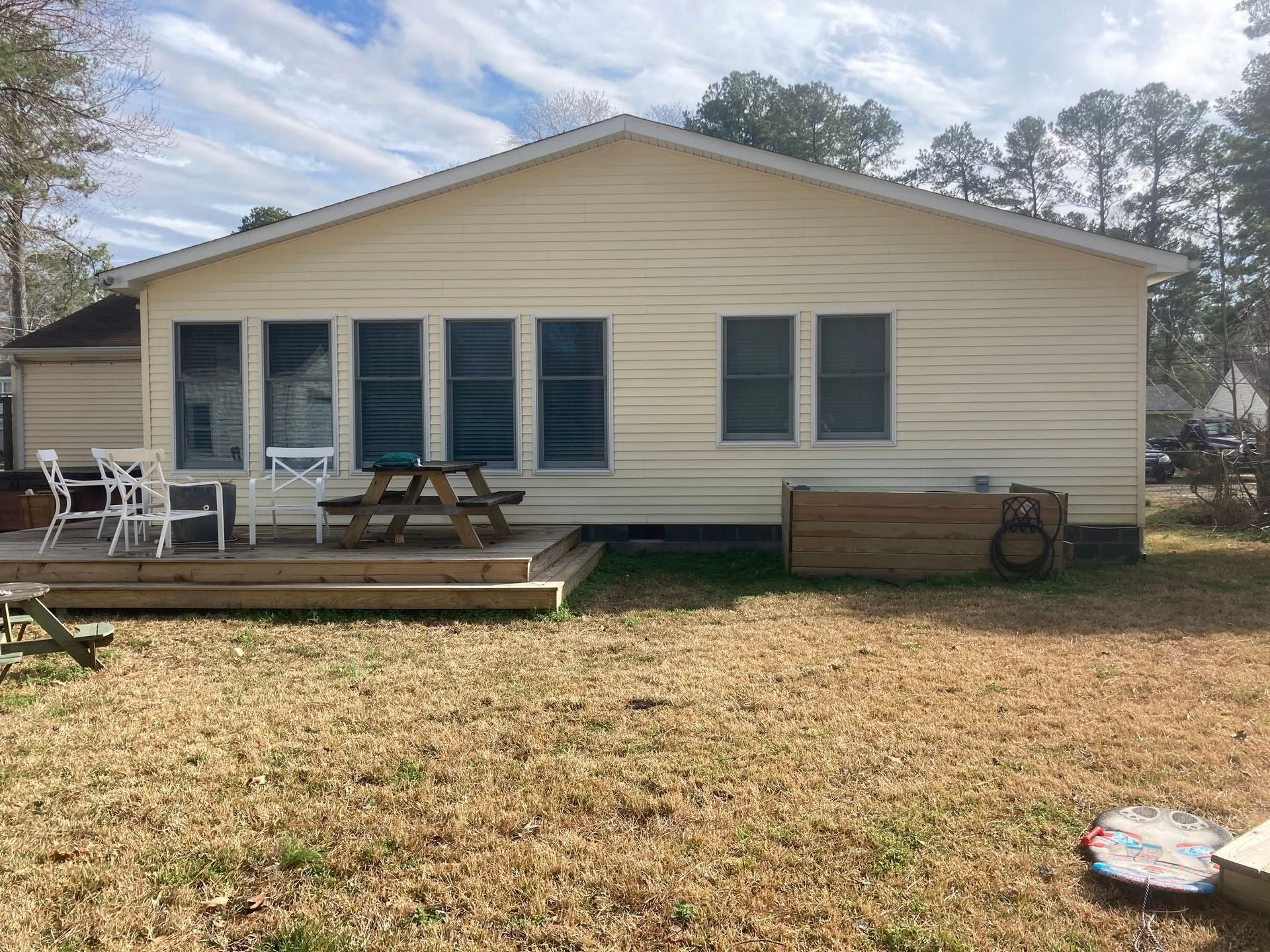 Single-story beige house with blue shutters, porch chairs, picnic table, and grassy yard under a cloudy sky