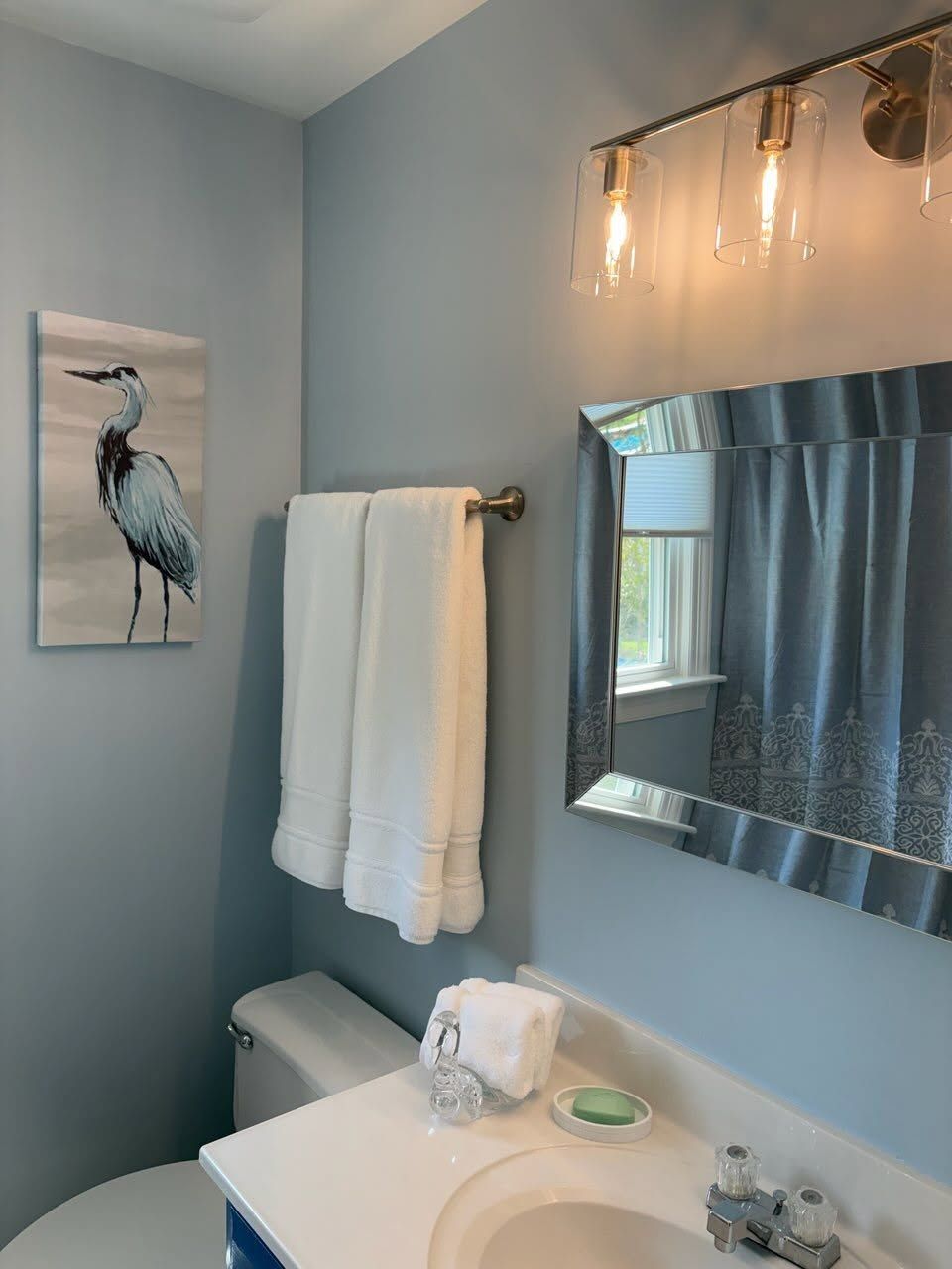 Blue bathroom with towel rack, flamingo wall art, mirror, sink, and shower curtain.