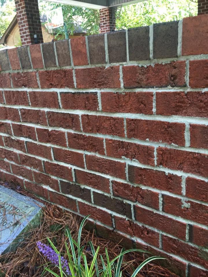 Red brick wall with white mortar, topped by darker bricks, beside grass and a purple flower in the foreground