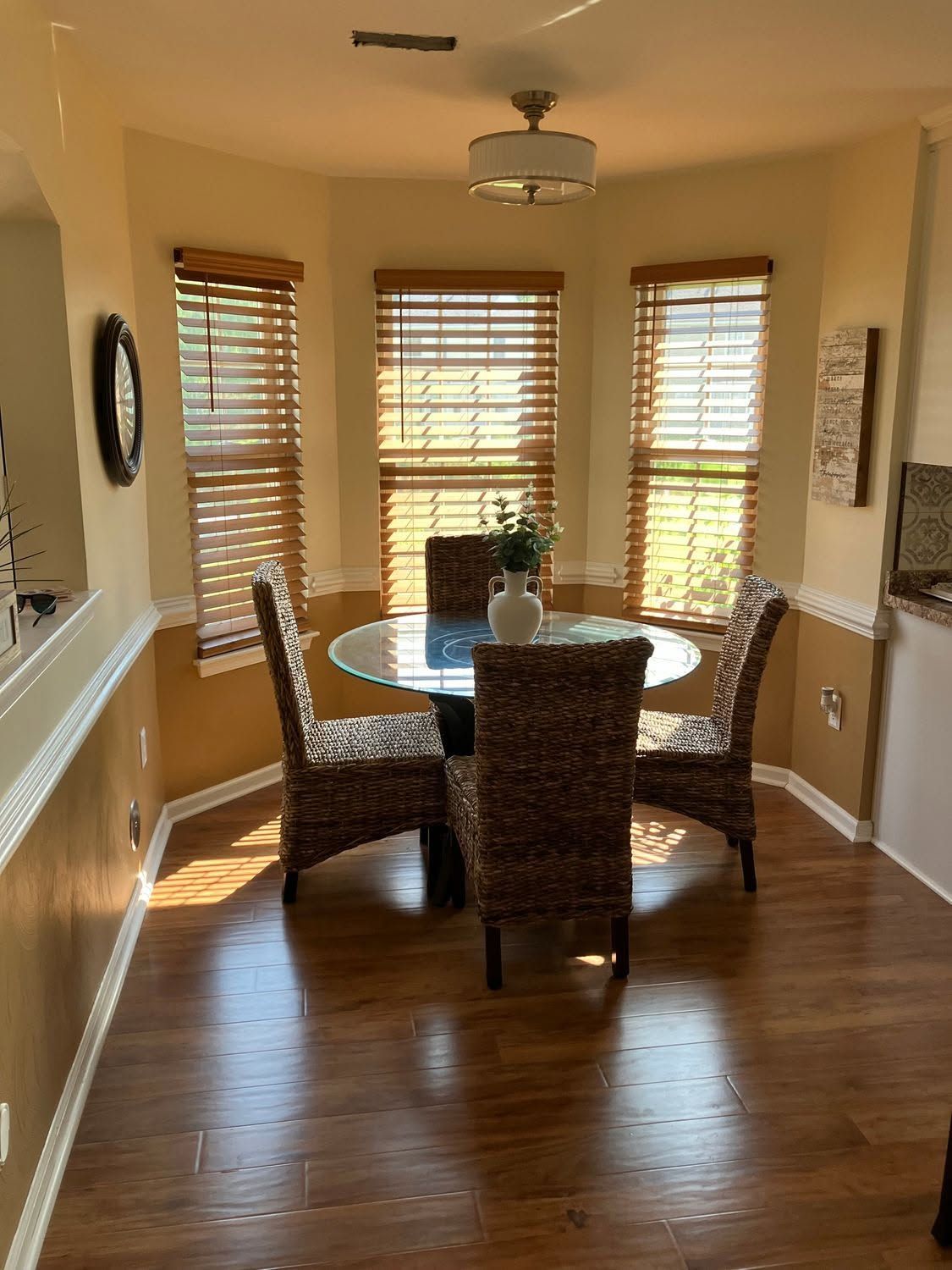 Bright dining room with round glass table, wicker chairs, wood floor, and sunlit windows.