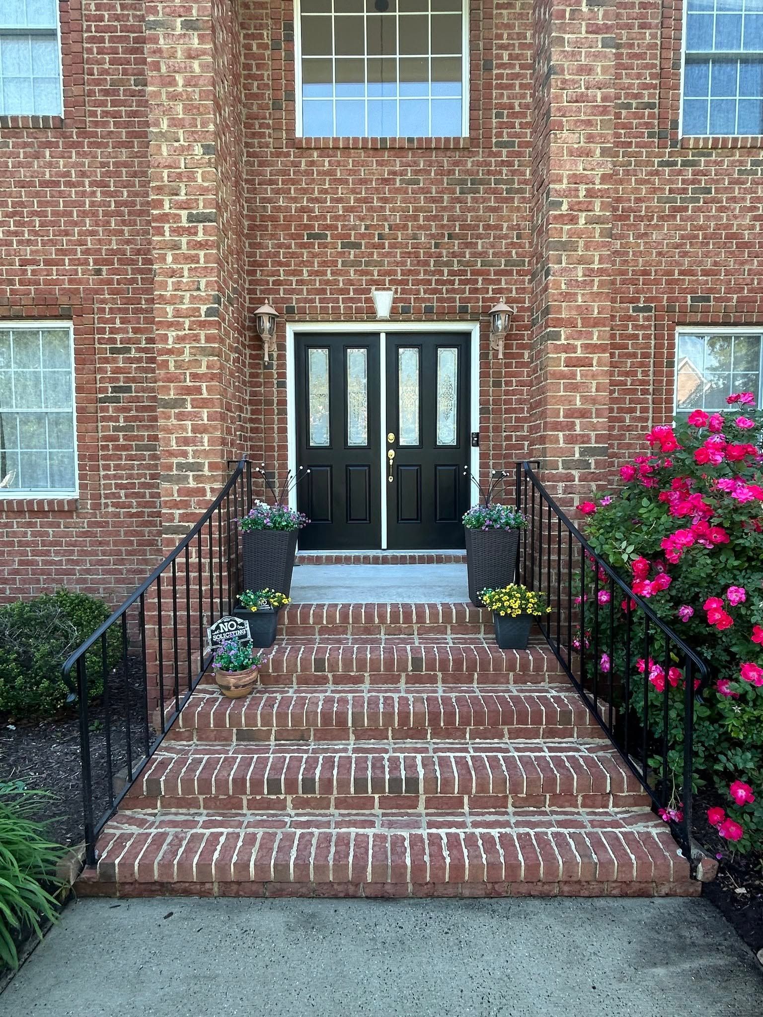 Brick townhouse entrance with black double doors, brick steps, and flower beds on both sides