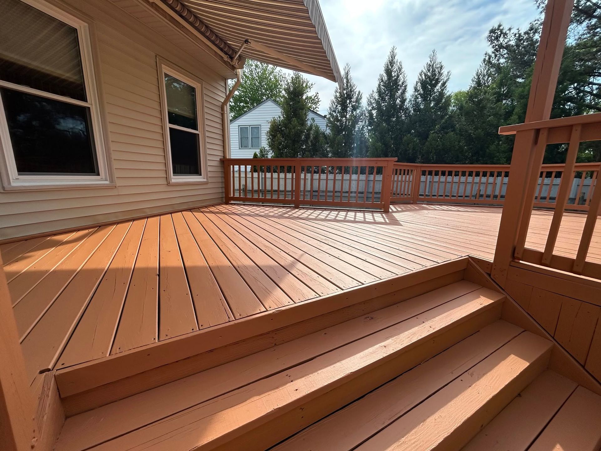 Wooden deck with steps, railing, and house siding beside a yard with trees