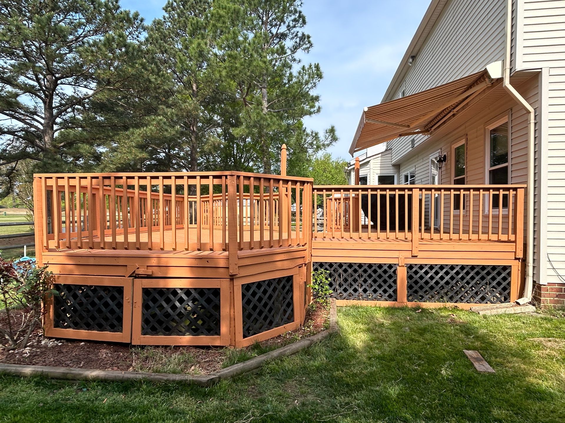 Wooden raised deck with railing and lattice skirting beside a brick house in a grassy yard