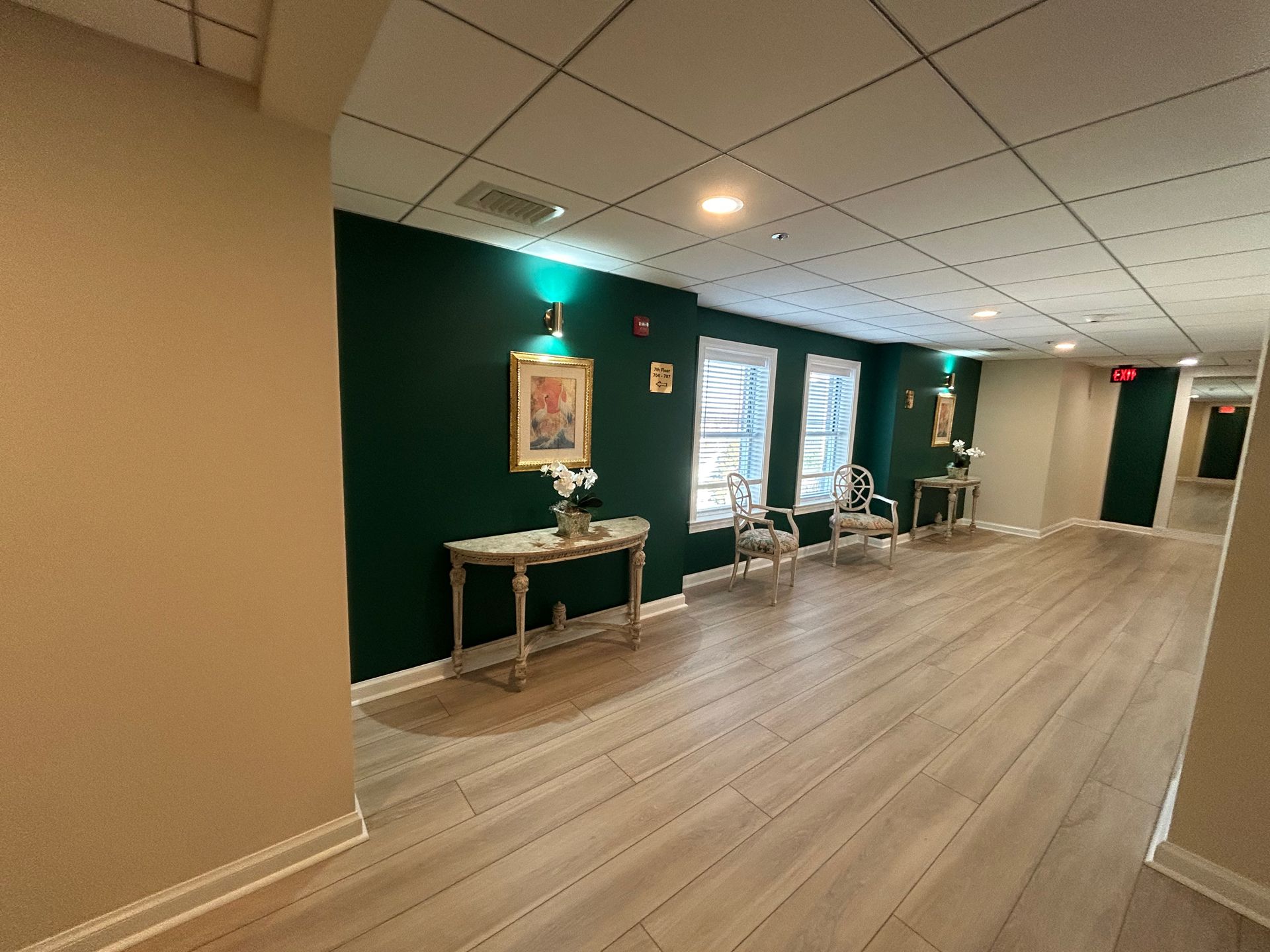 Empty hallway with green accent wall, framed art, chairs, and a table under bright ceiling lights