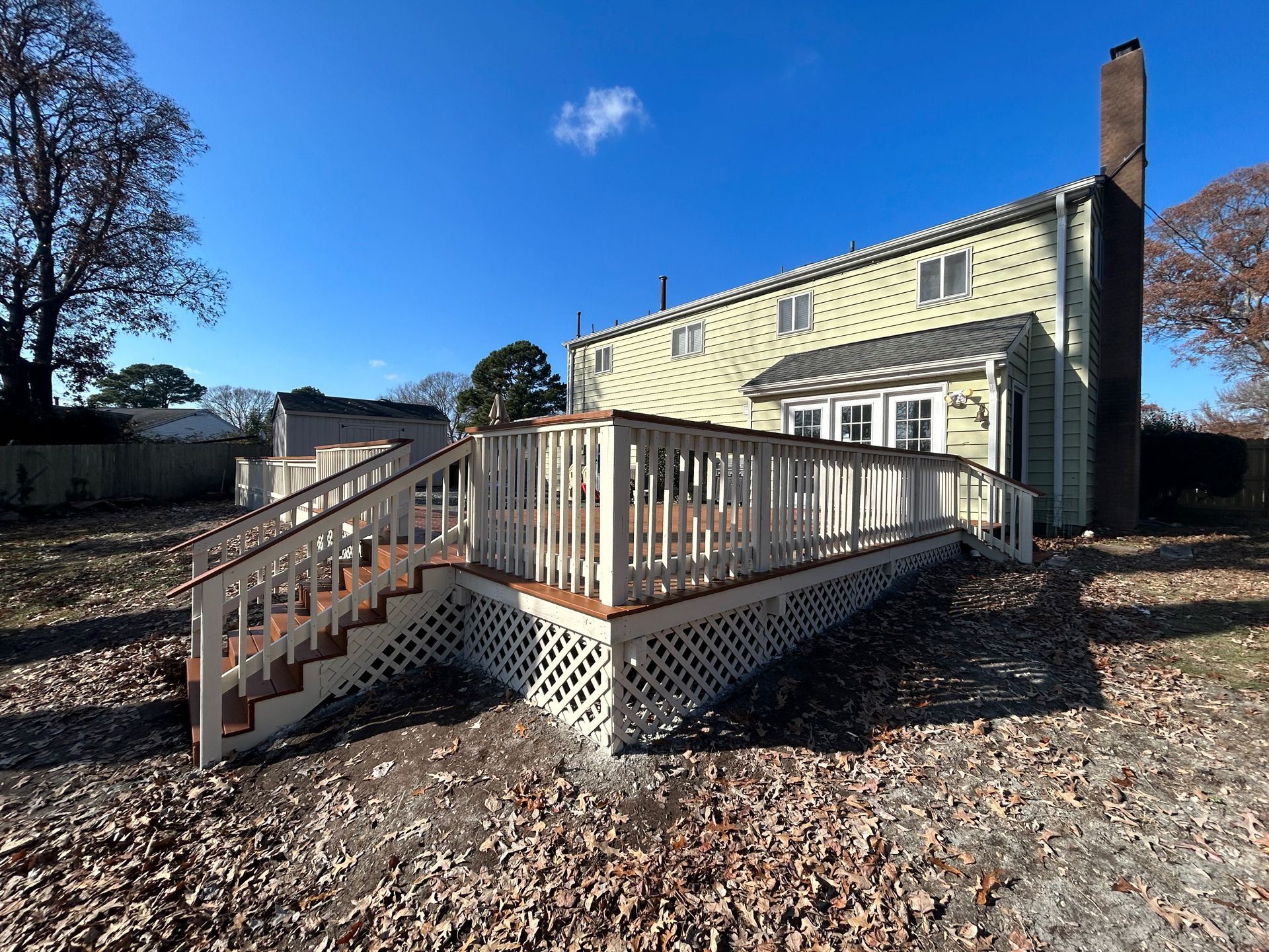 Back deck with white railing on a yellow house, set in a leaf-covered yard under a blue sky