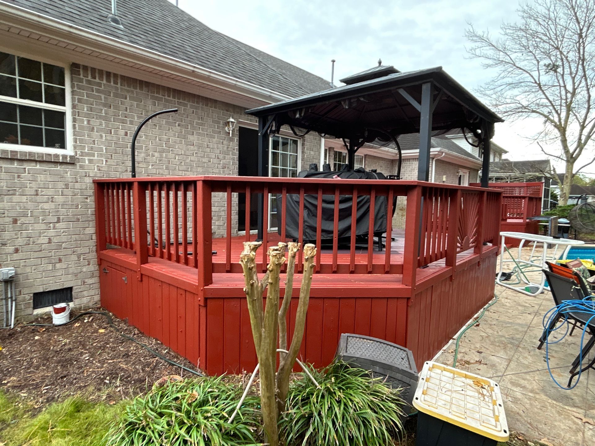 Red wooden deck with railings attached to a brick house, with patio furniture and a pergola.