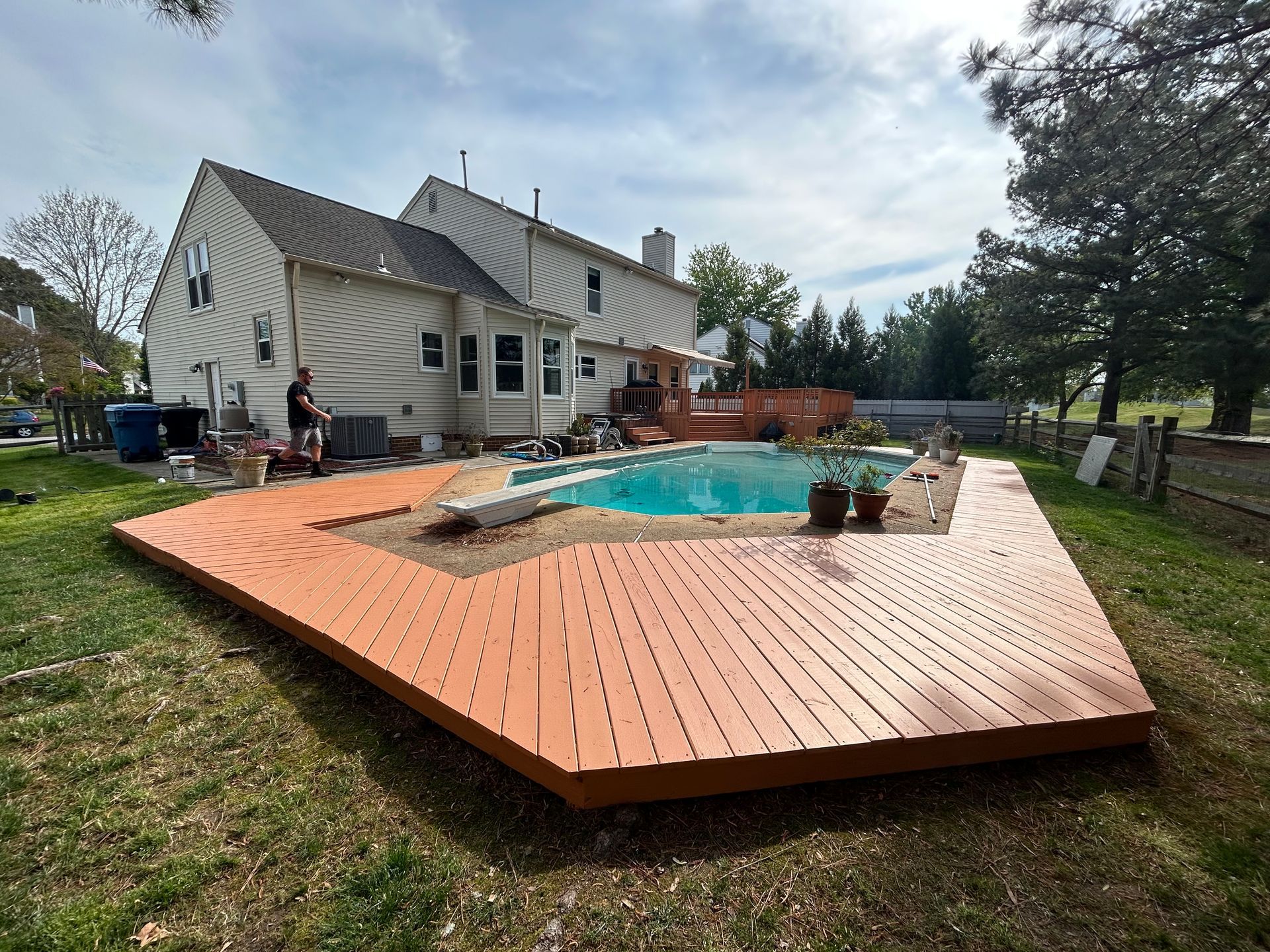 Backyard deck around an above-ground pool beside a white house, with trees and lawn nearby