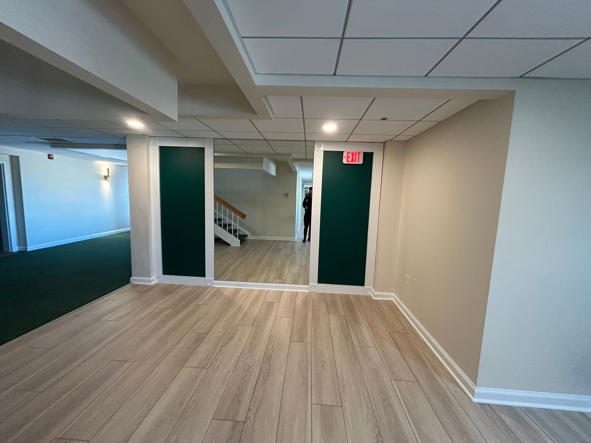 Empty basement room with light wood flooring, white walls, teal doors, and an EXIT sign.
