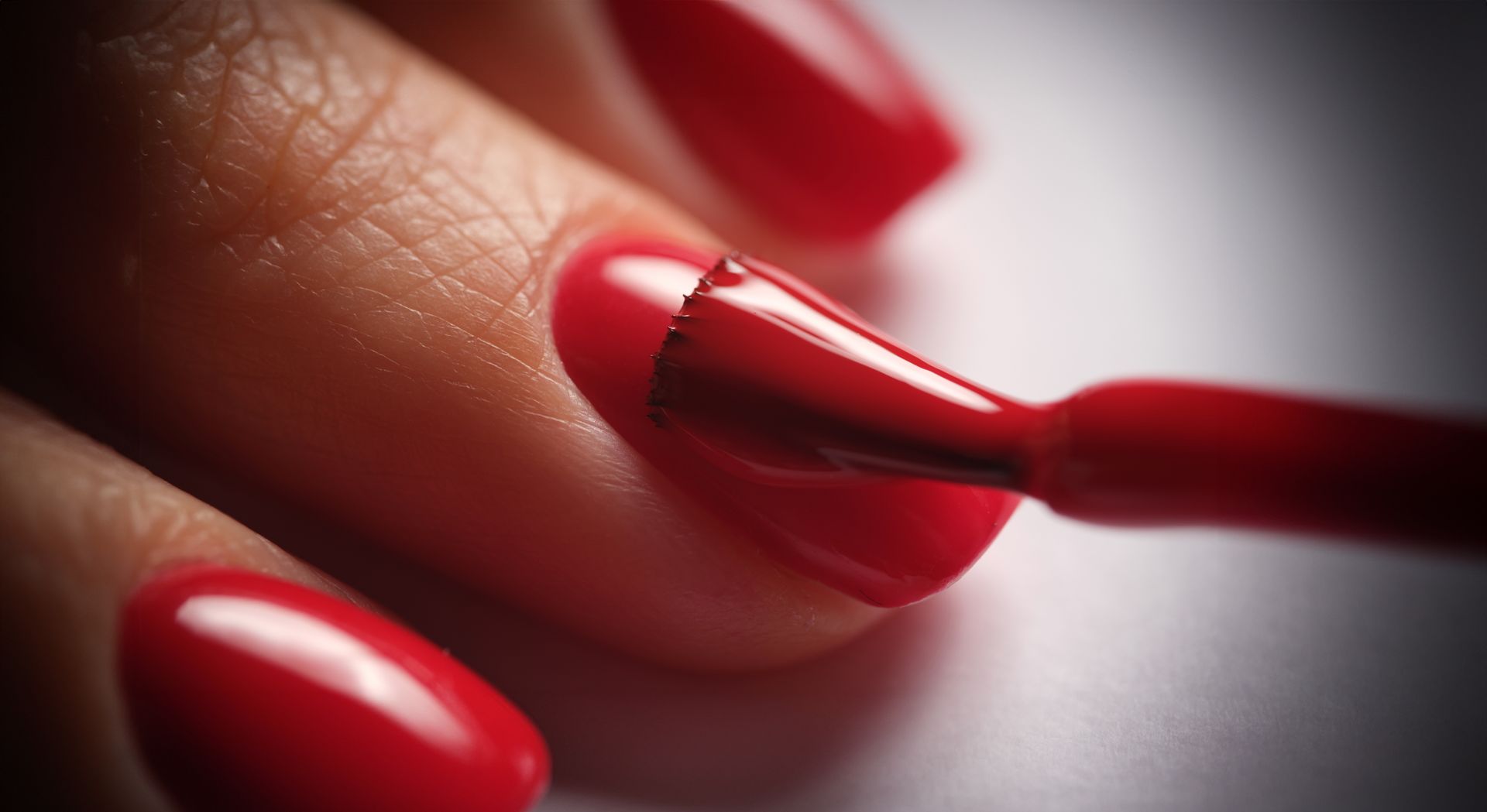 Un pinceau applique du vernis à ongles rouge brillant sur un ongle, un autre ongle fraîchement verni en rouge étant visible à proximité.