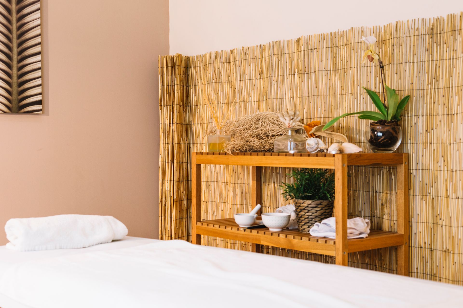 Une table de massage avec une serviette blanche à côté d'une étagère en bambou contenant des produits de spa et une plante en pot, adossée à un mur de roseaux.