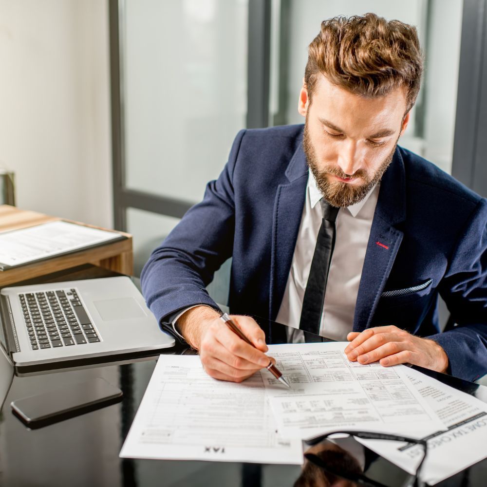 Man in navy suit reviewing papers at an office desk beside an open laptop