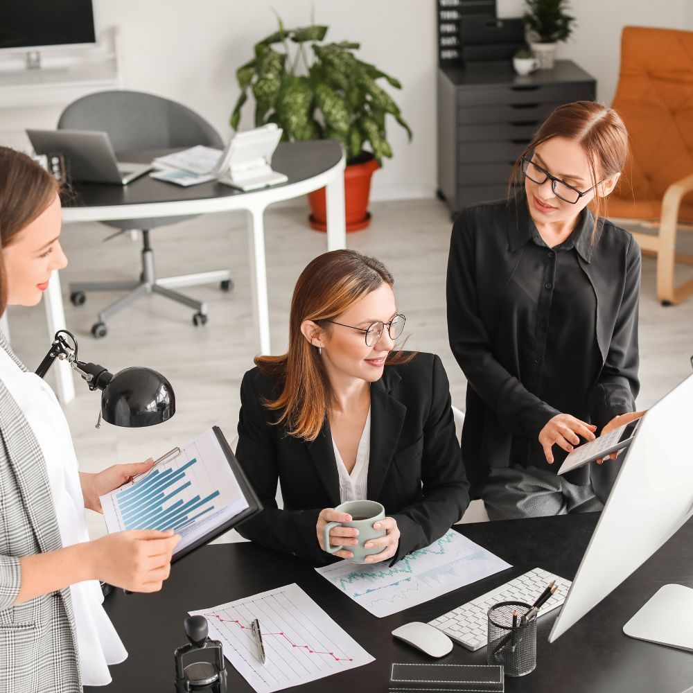 Three coworkers in a bright office discussing documents around a desk with a computer.