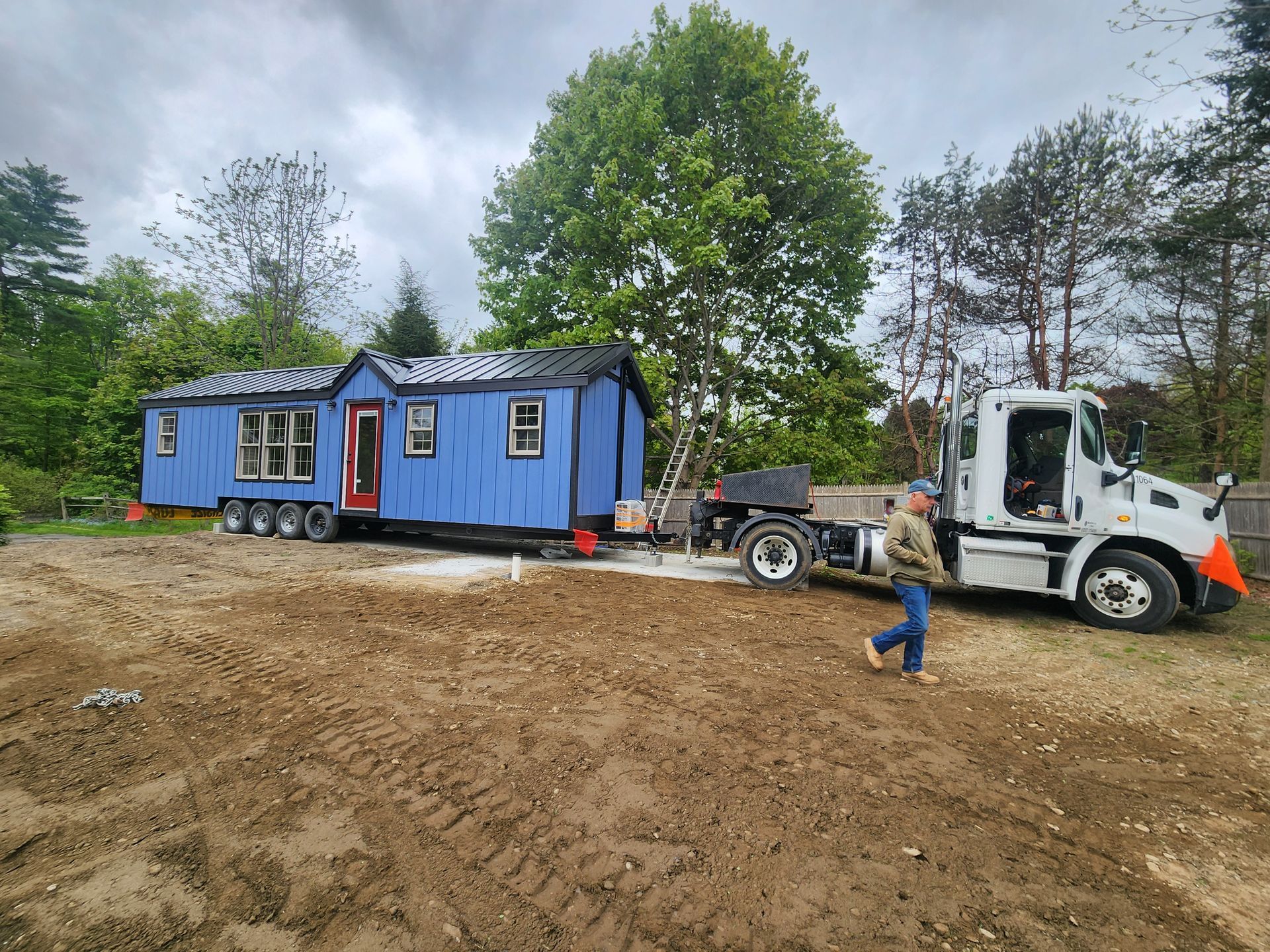 A blue modular house is being delivered on a semi-truck trailer across a dirt lot, with a worker standing nearby.