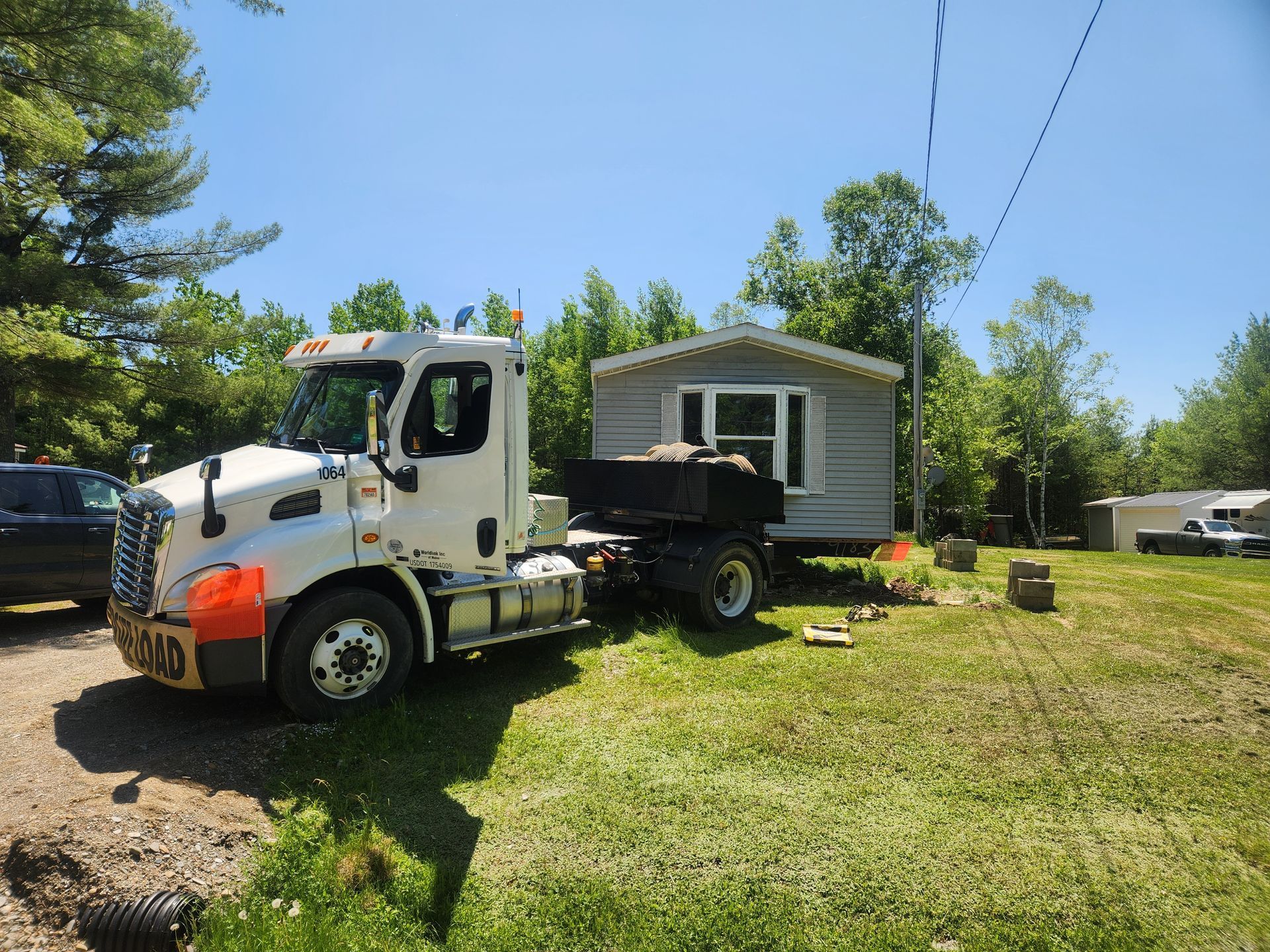 A white semi-truck parked in a grassy area, hitched to a gray prefabricated home being moved on a trailer.