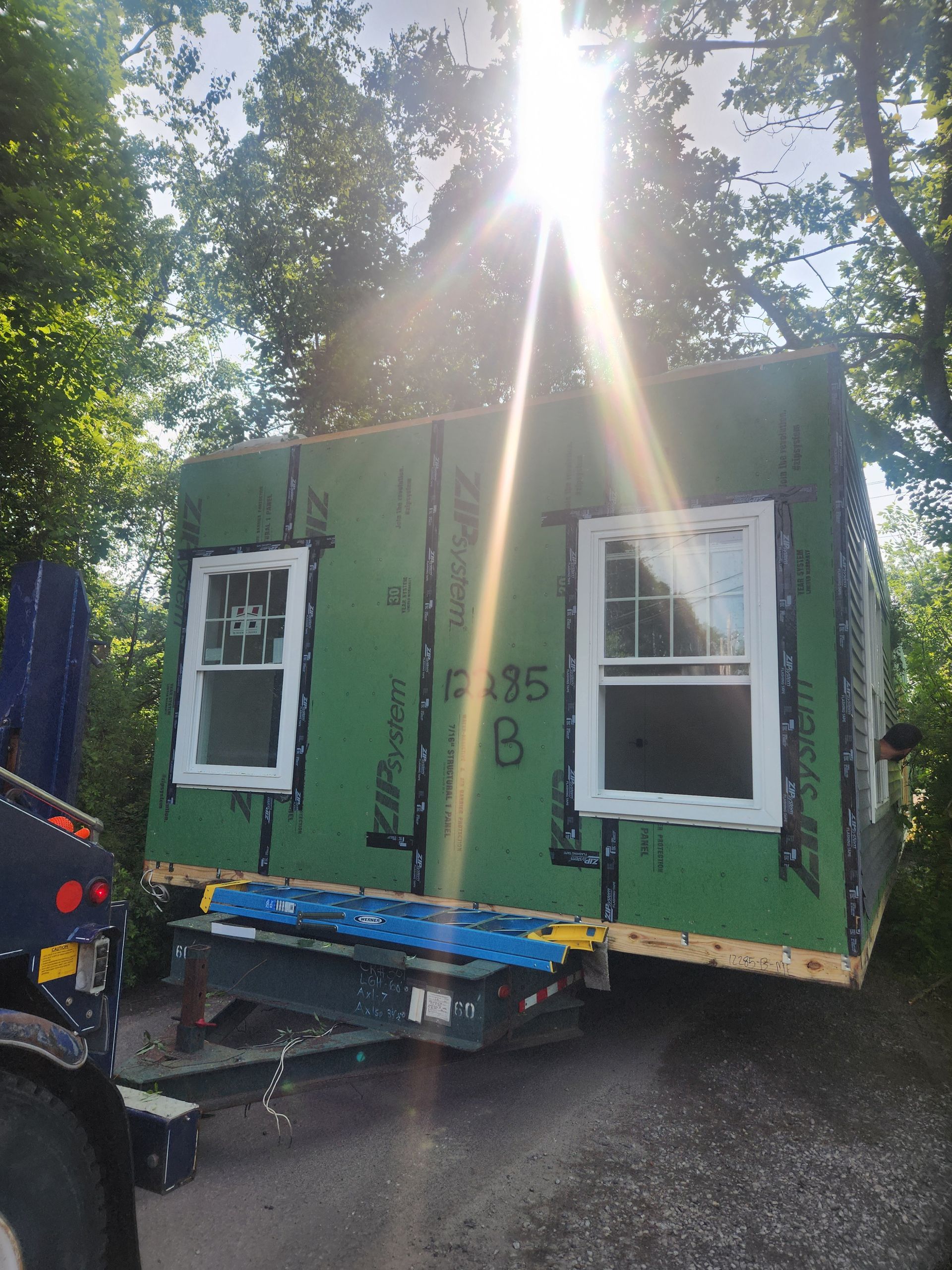 A small house frame on a trailer, featuring green plywood walls with two white windows, parked outdoors under sunlight.