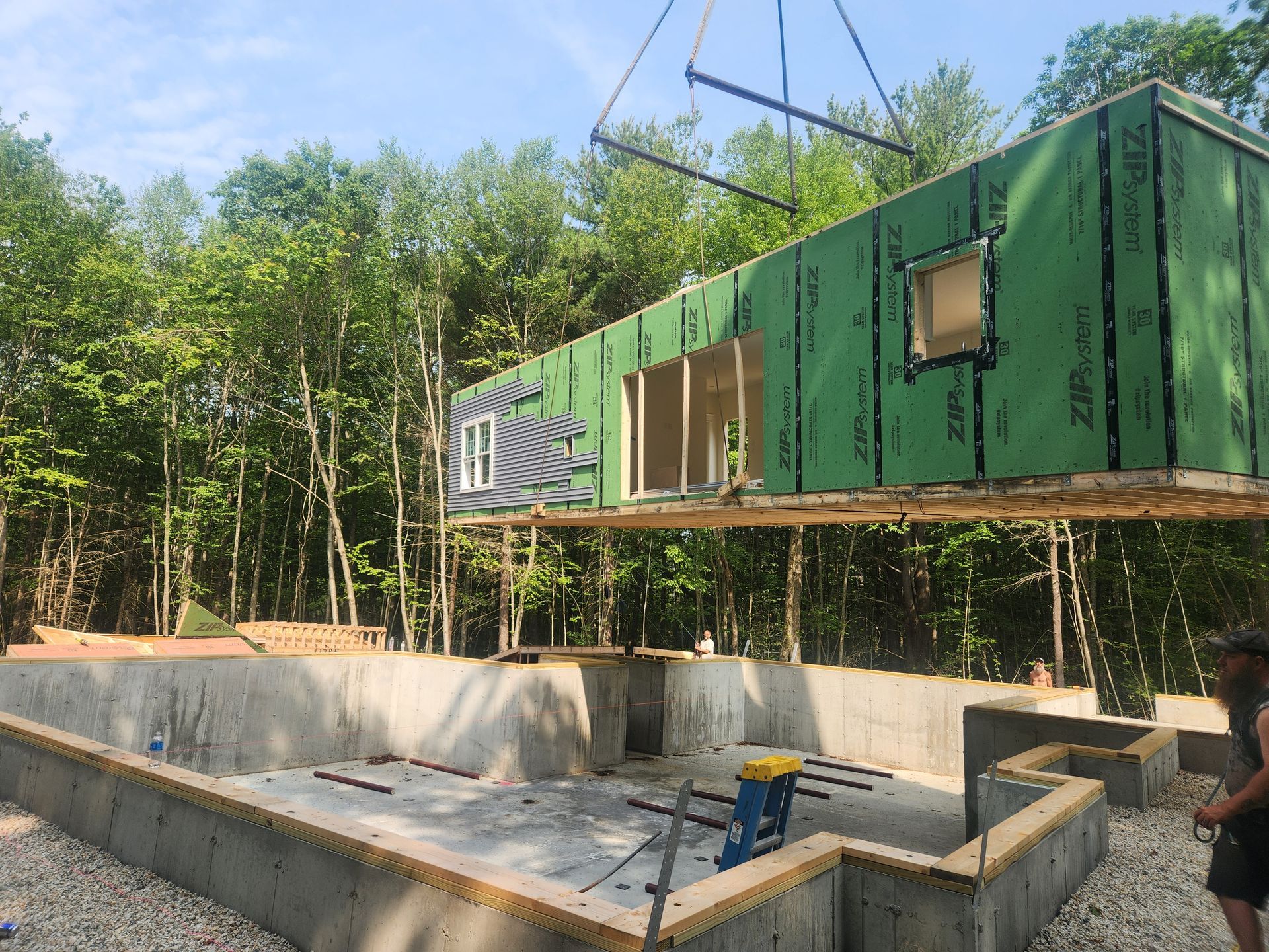A crane lowers a green modular home section onto a concrete foundation in a wooded setting.