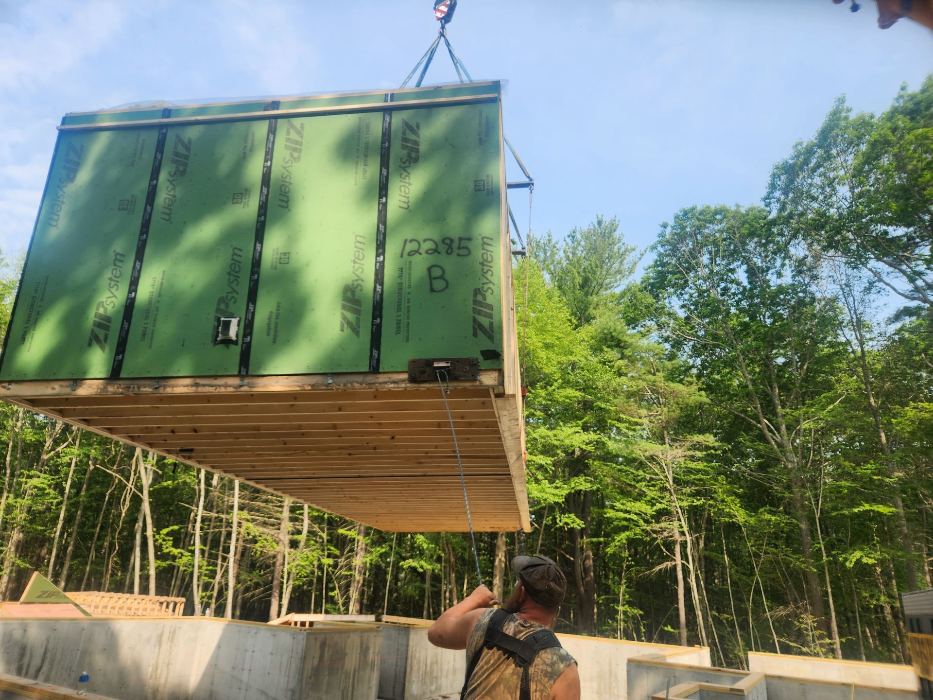 A large modular house section is suspended by a crane over a concrete foundation in a wooded area.