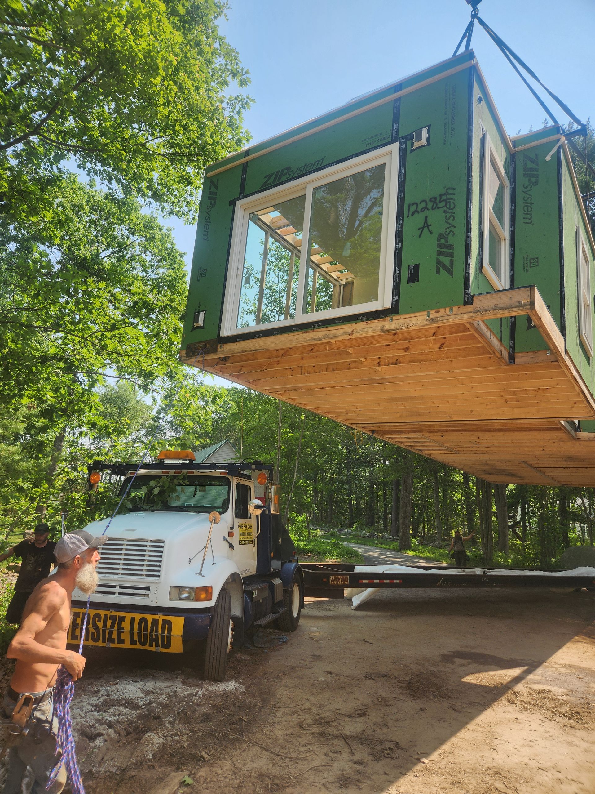 A crane lifts a modular home section over a truck in a wooded area while a worker holds a guide rope.