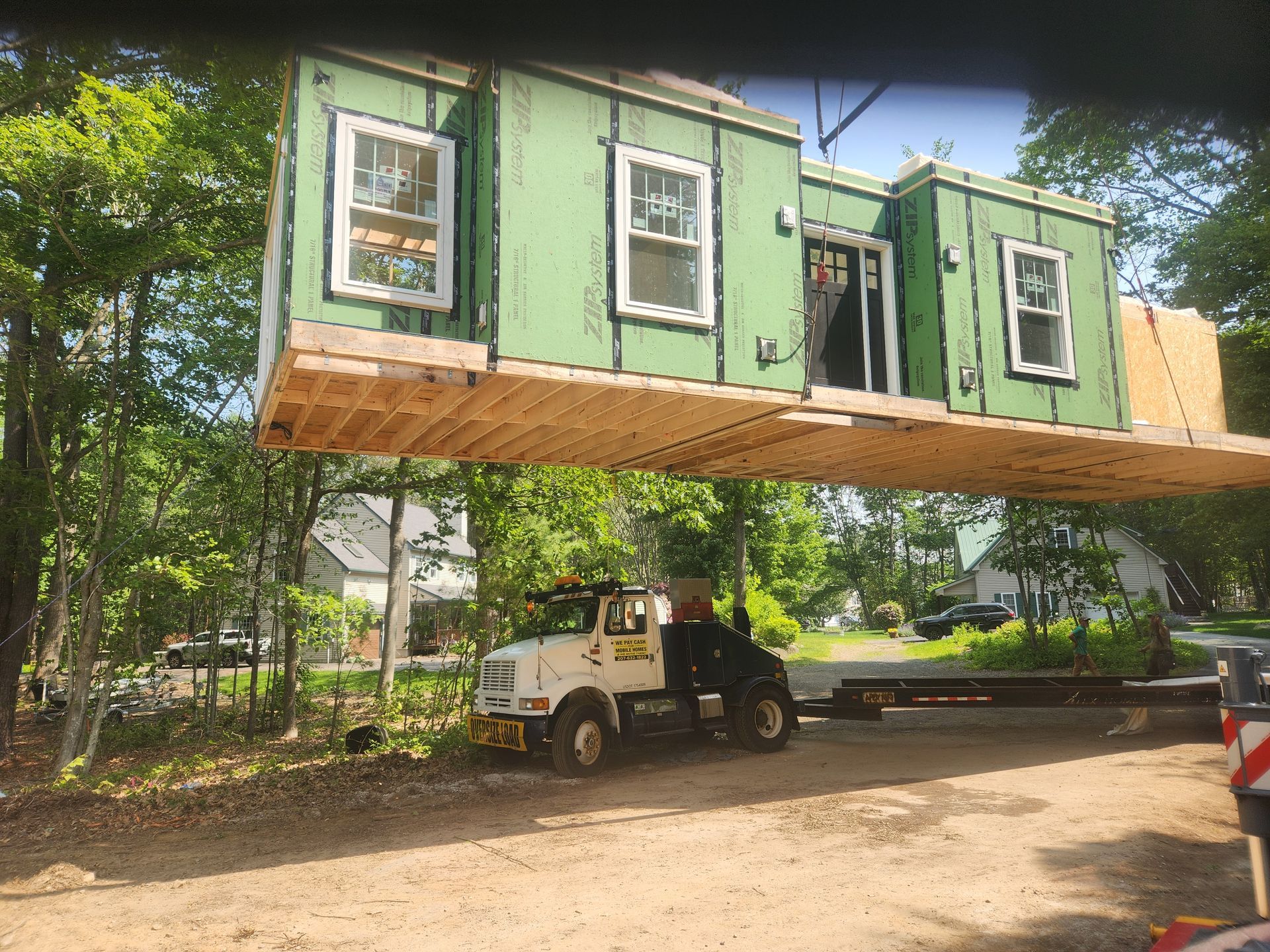 A crane lifts a modular house section, framed with green sheathing and white-trimmed windows, over a truck in a wooded area.