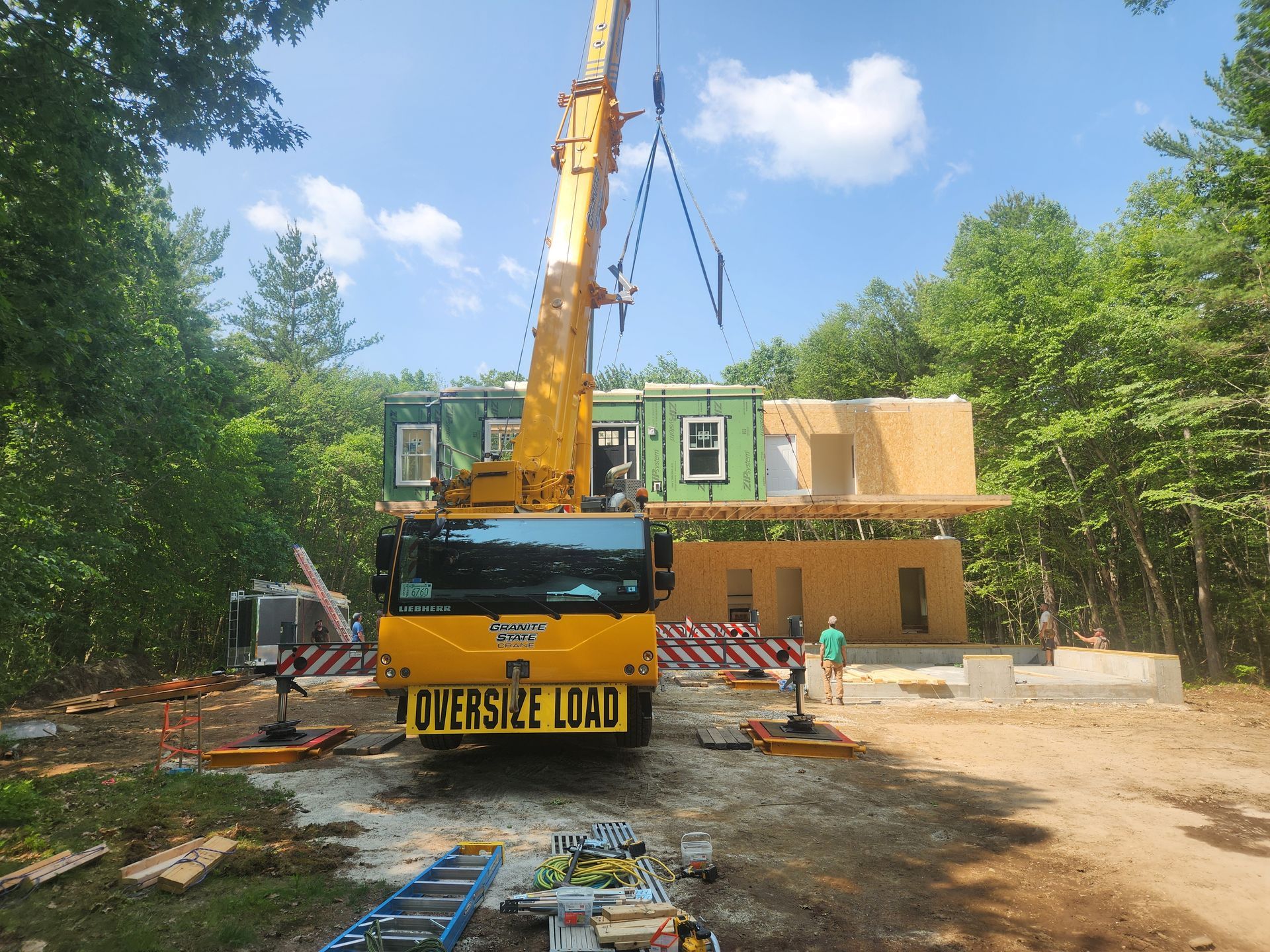 A large yellow mobile crane lifting a modular home section onto a wooded residential construction site.