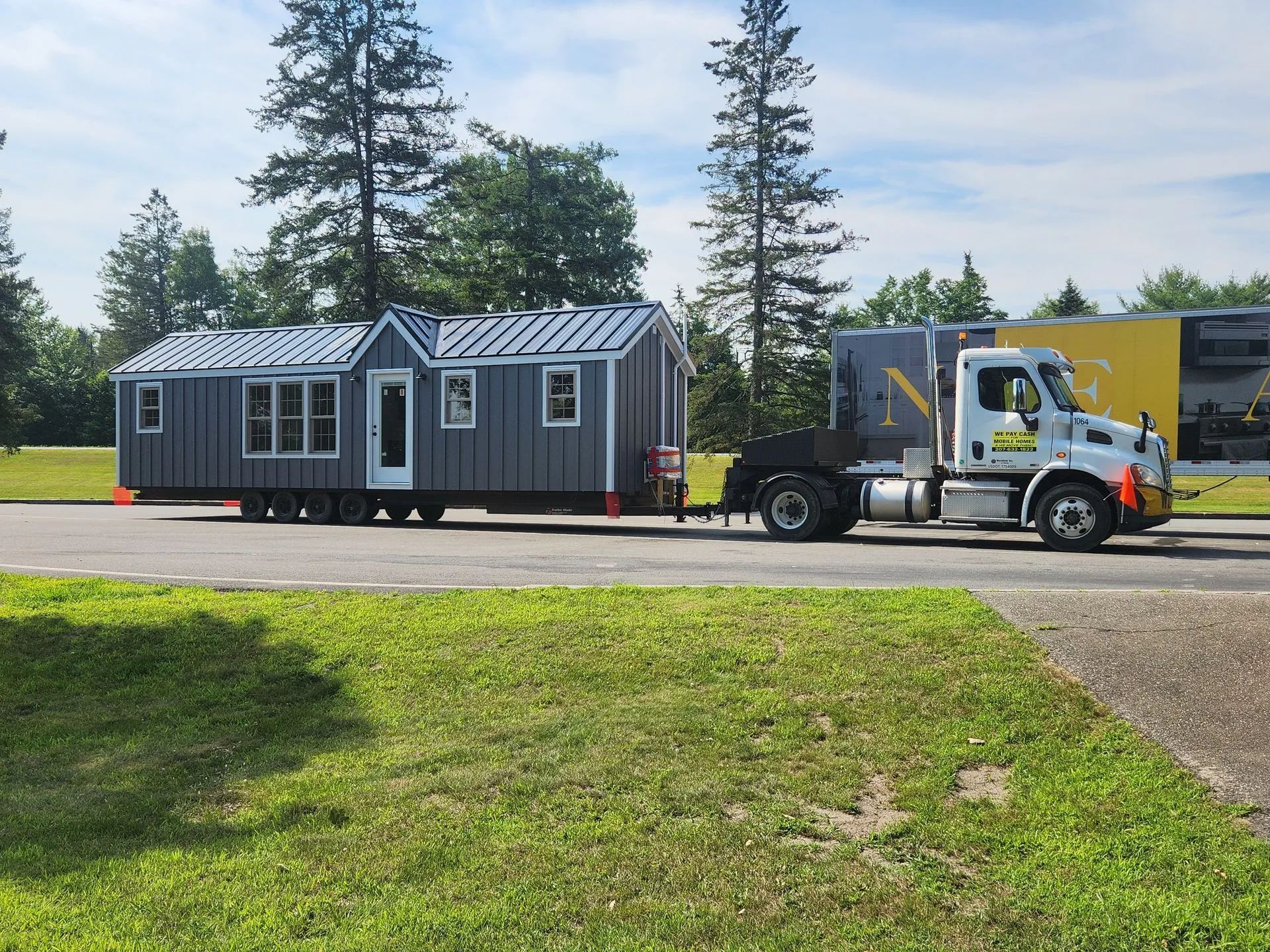 Tractor hauling a gray tiny house on a trailer along a road near a grassy field and trees