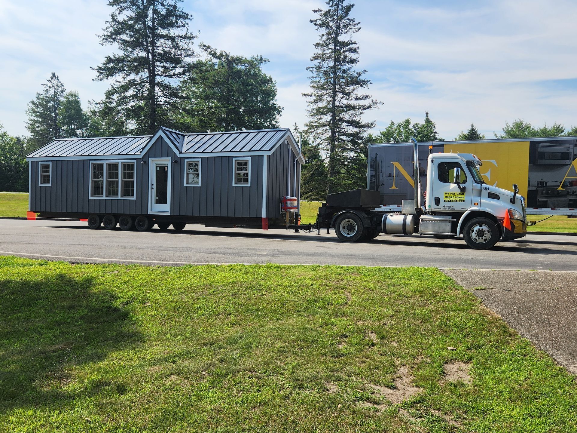 A white semi-truck pulling a grey tiny home on a trailer along a paved road surrounded by trees and grass.