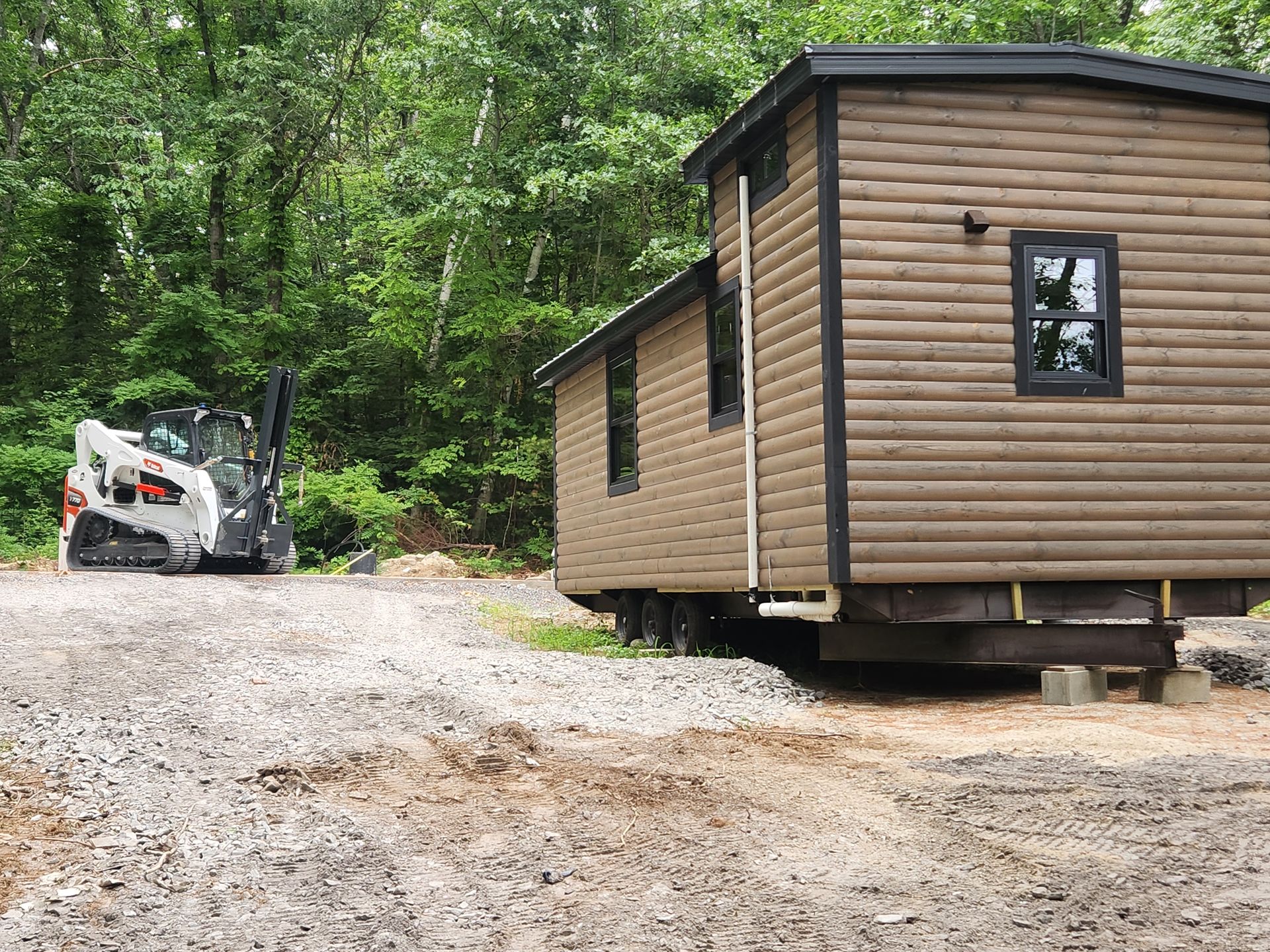 A brown tiny house on a trailer sits on a gravel lot next to a small white skid steer loader near a wooded area.