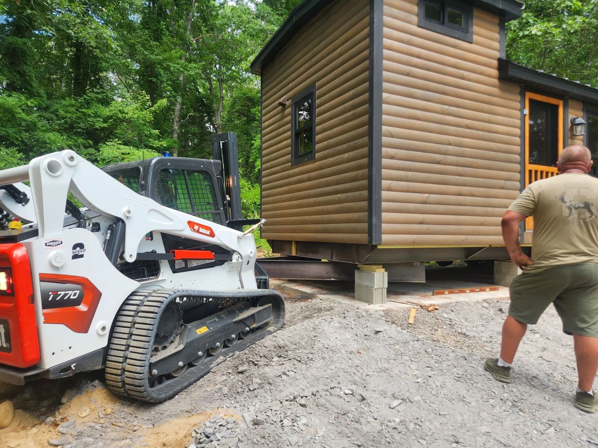 Skid steer moving a small log cabin on blocks while a man guides it in a wooded yard