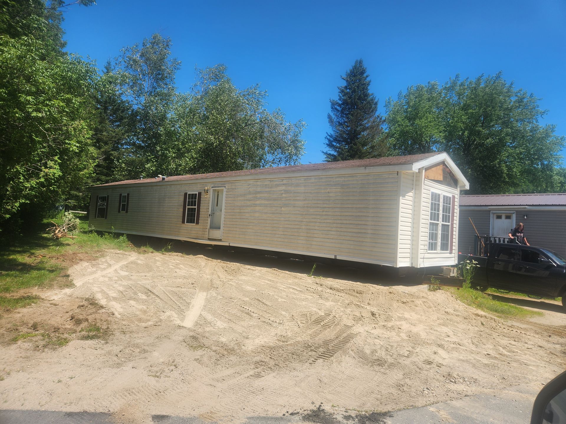 A long, beige manufactured home sits on a dirt slope under a clear blue sky, with trees and a truck visible nearby.