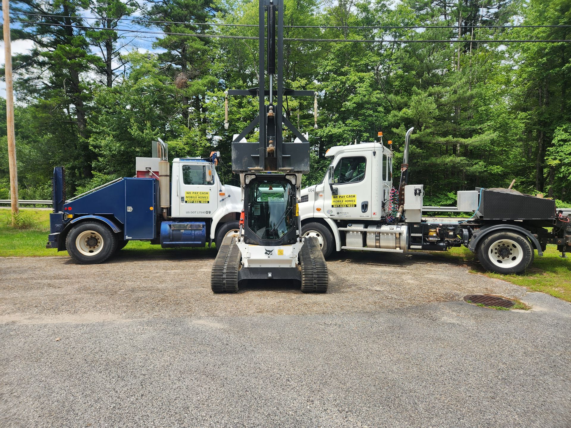 Two white semi-trucks parked on a gravel lot with a tracked forestry machine positioned between them.