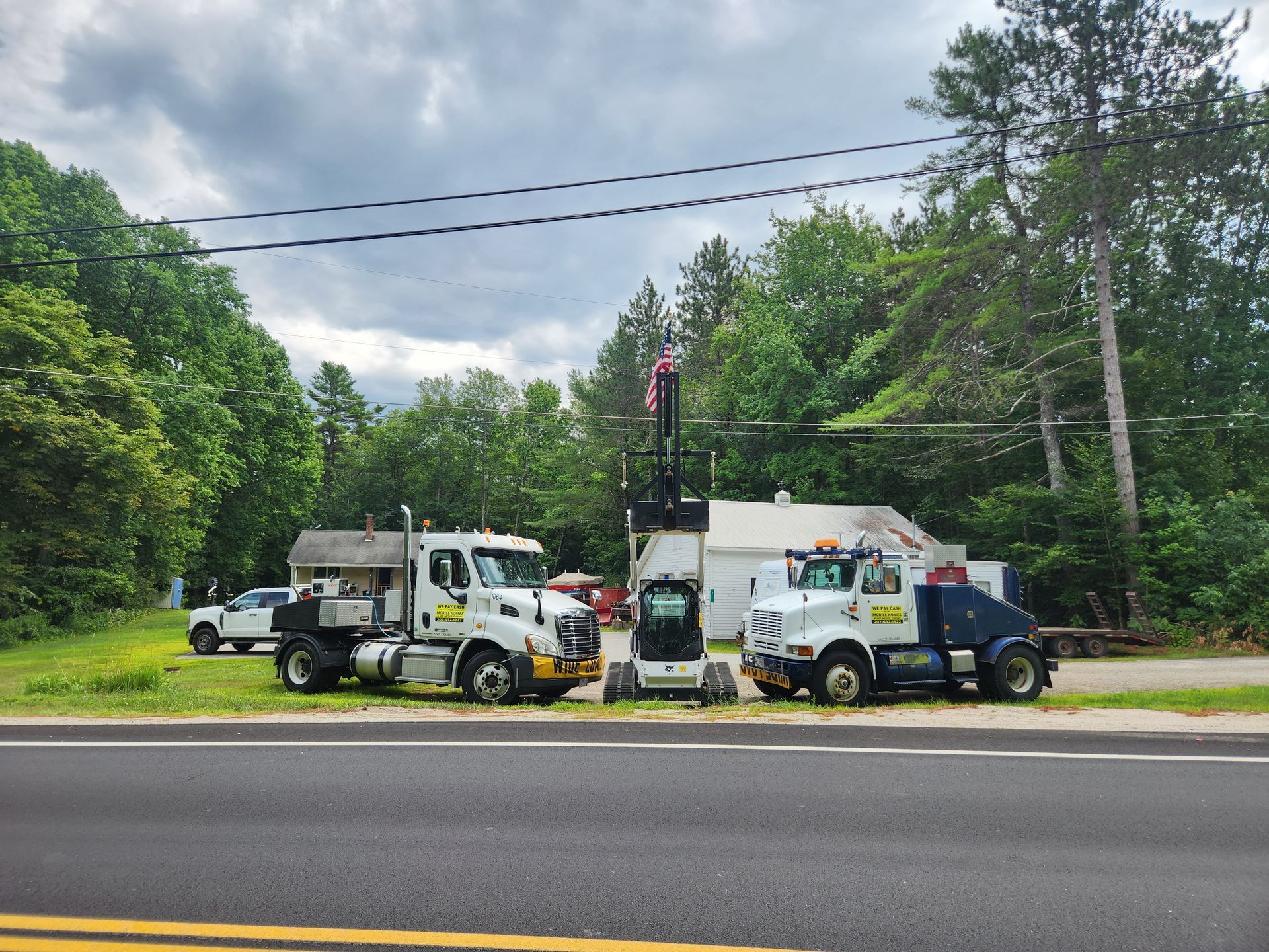 Two white work trucks and a smaller construction vehicle parked on a roadside lot with trees and a building in the back.