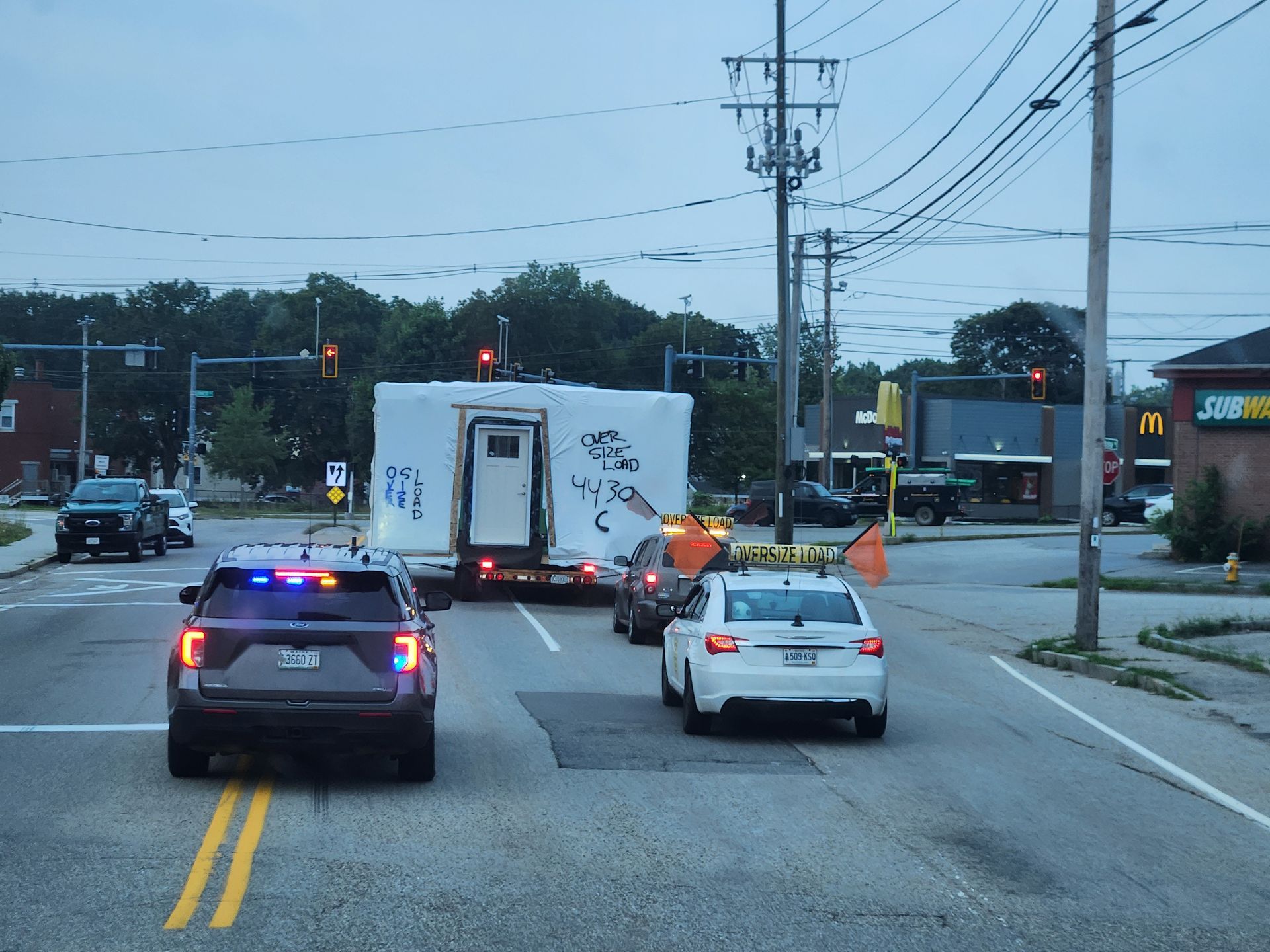 A police SUV with flashing lights leads a wide-load trailer through an intersection past a Subway restaurant.