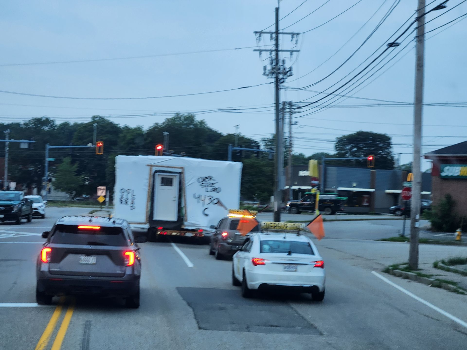 A white portable building is being towed through an intersection by a pilot car with an orange strobe light on its roof.