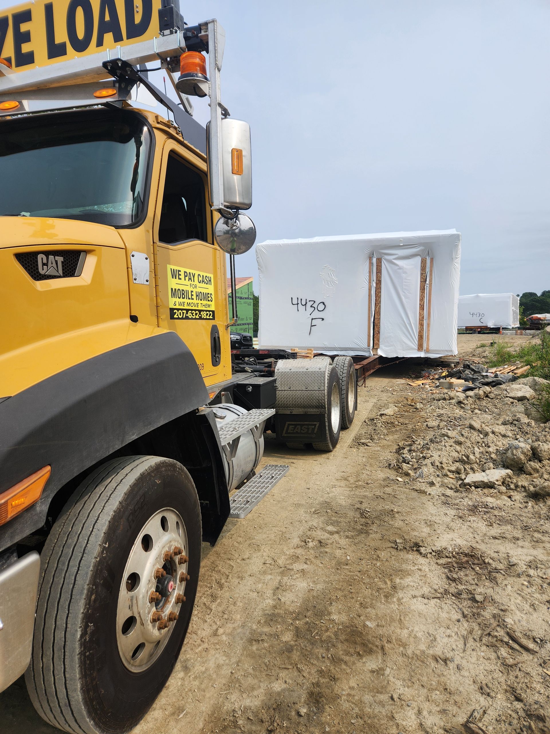 A yellow semi-truck pulls a large, white, shrink-wrapped modular structure on a trailer over a gravel site.