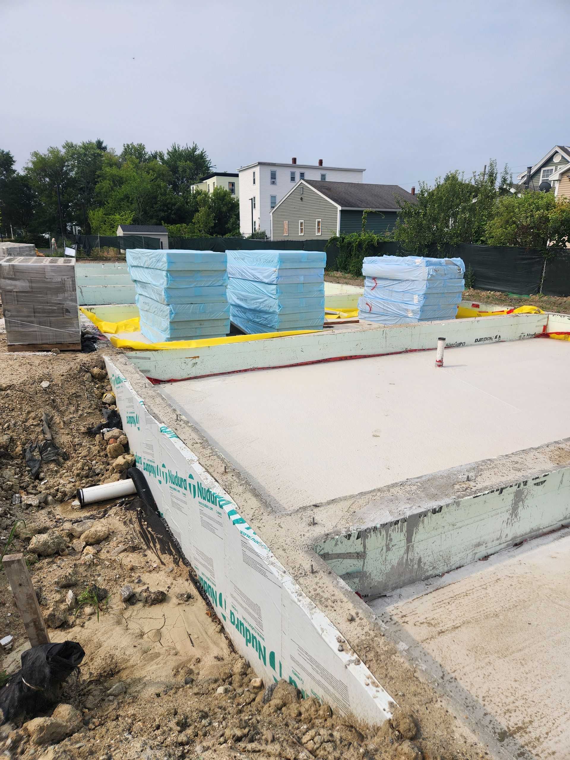 Construction site showing a concrete foundation, stacks of blue rigid foam insulation, and yellow vapor barrier.
