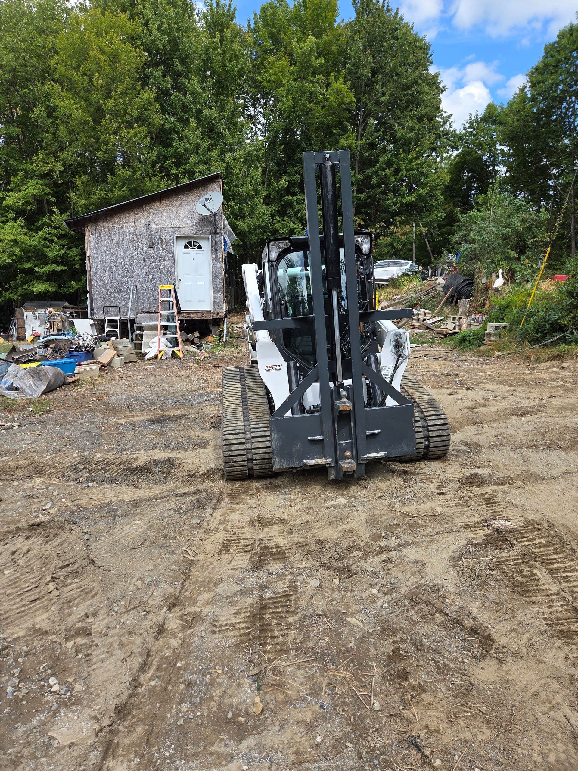 A white skid-steer loader with a black fork attachment parked on a dirt lot in front of a rustic, weathered cabin.