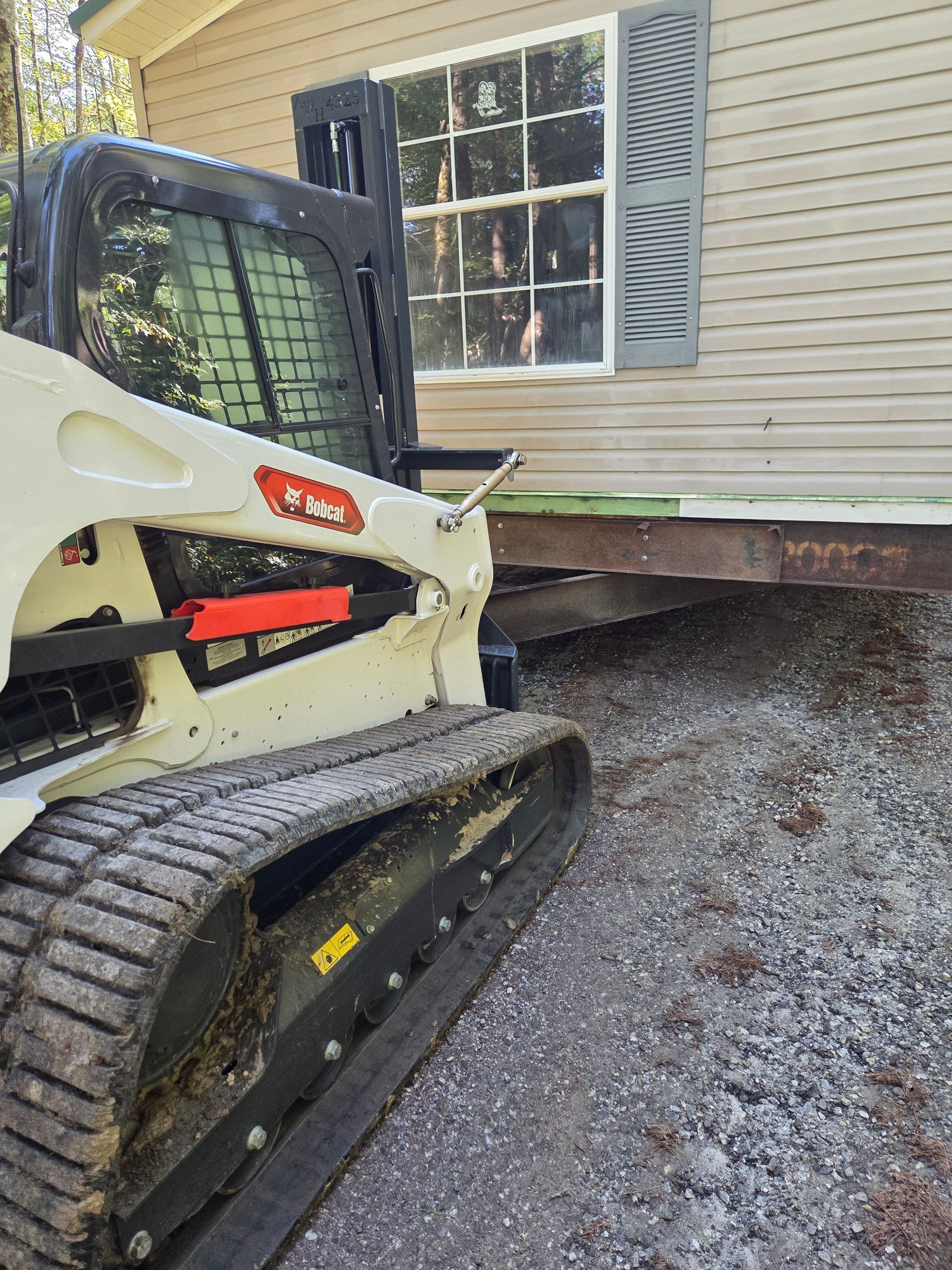 A white compact track loader parked next to a house structure resting on steel I-beams on a gravel surface.