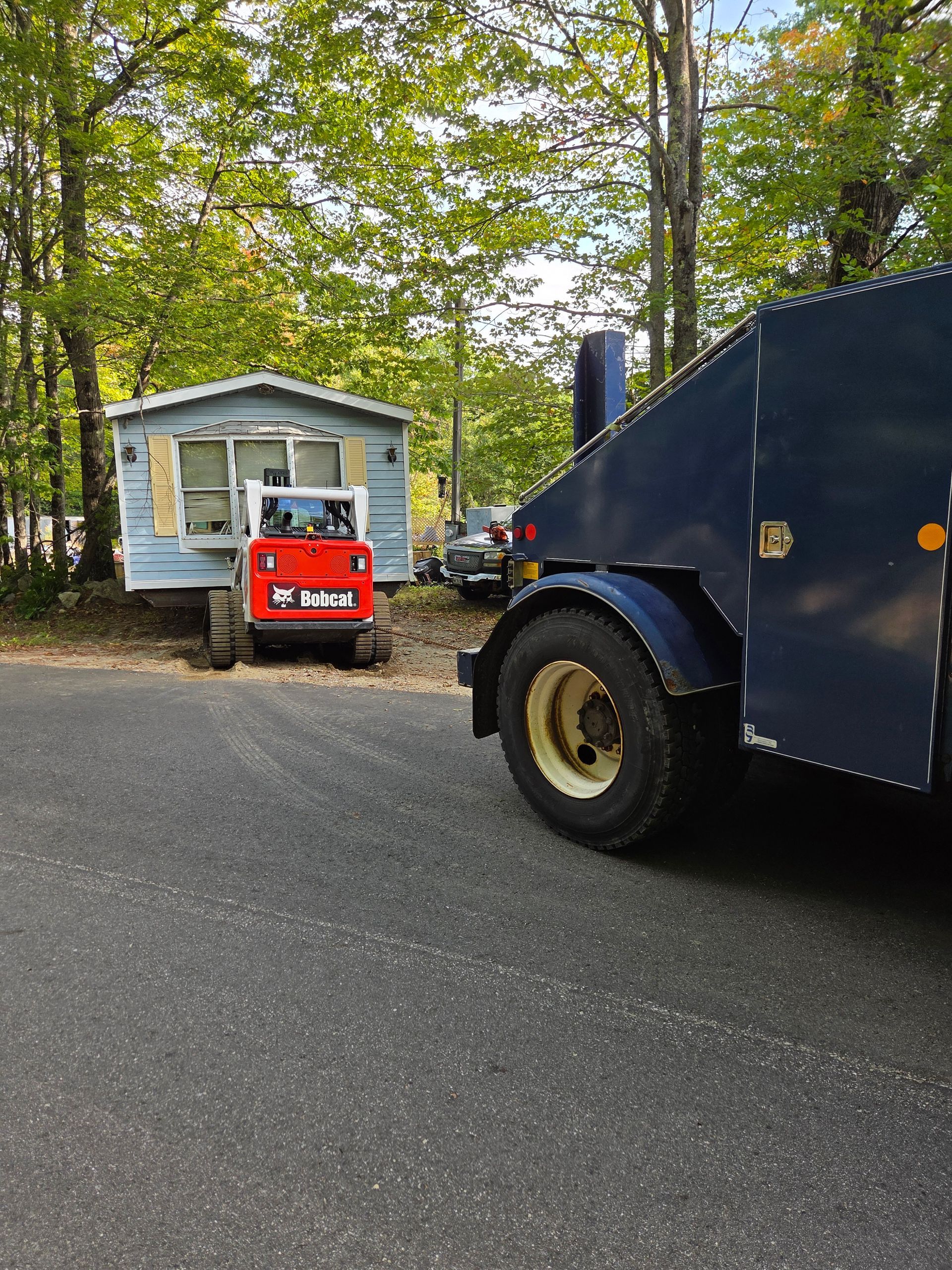 A red compact loader parks in front of a blue mobile home near a dark truck on a paved road in a wooded area.