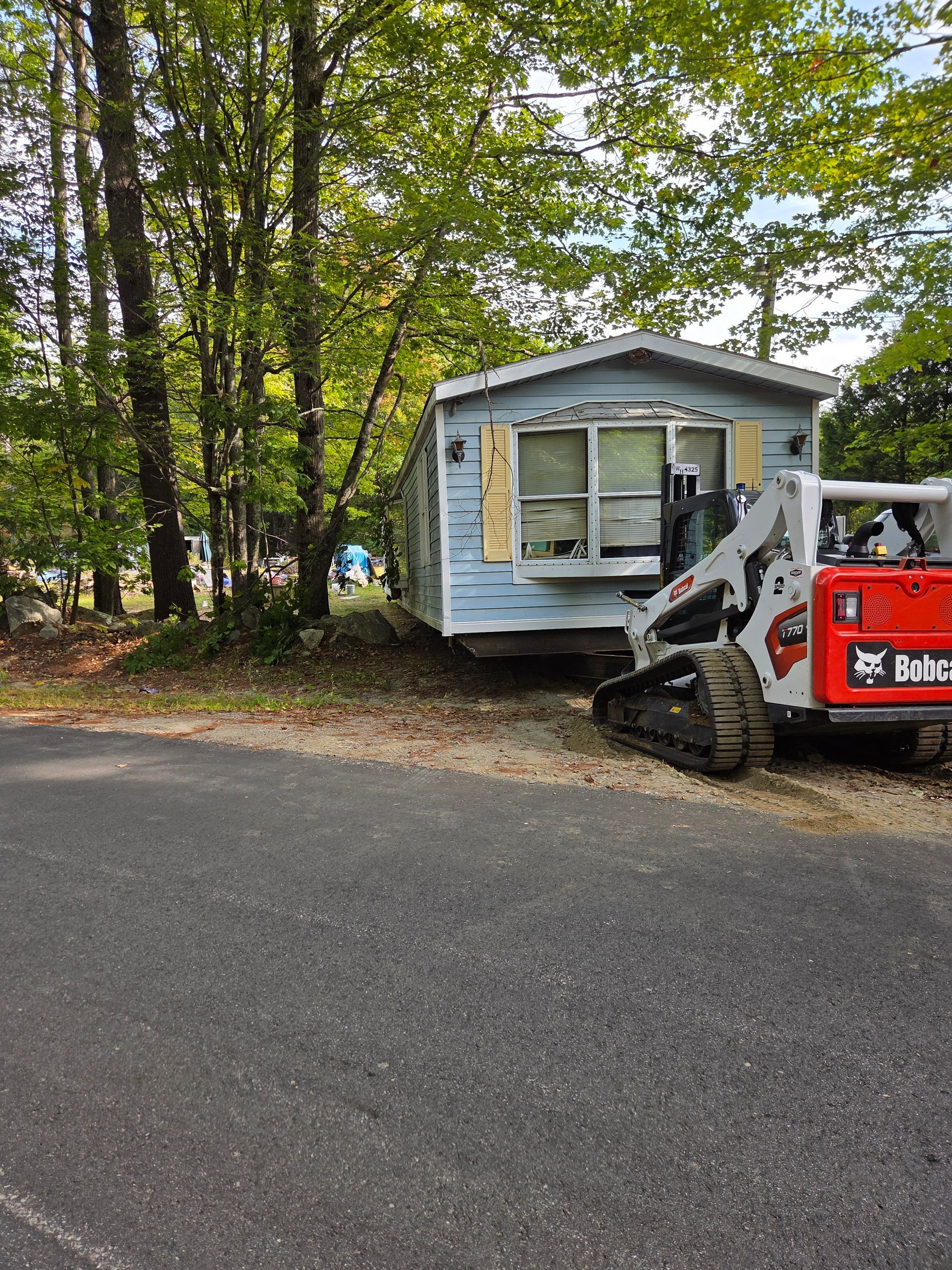 A red Bobcat track loader sits beside a light blue mobile home parked on a gravel lot near a wooded area.