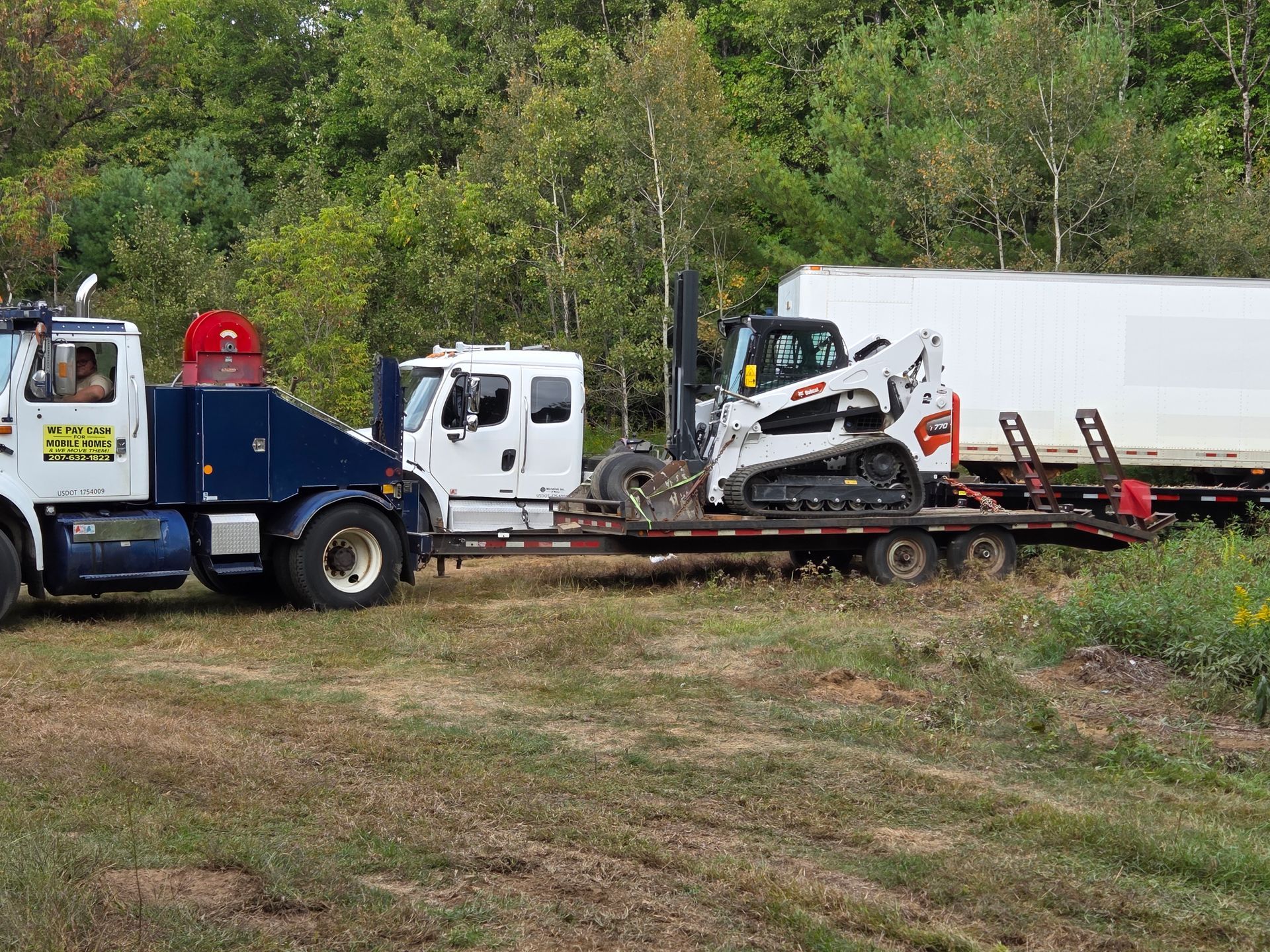 A white and blue tow truck pulls a flatbed trailer carrying a white Bobcat skid-steer in a field near a wooded area.
