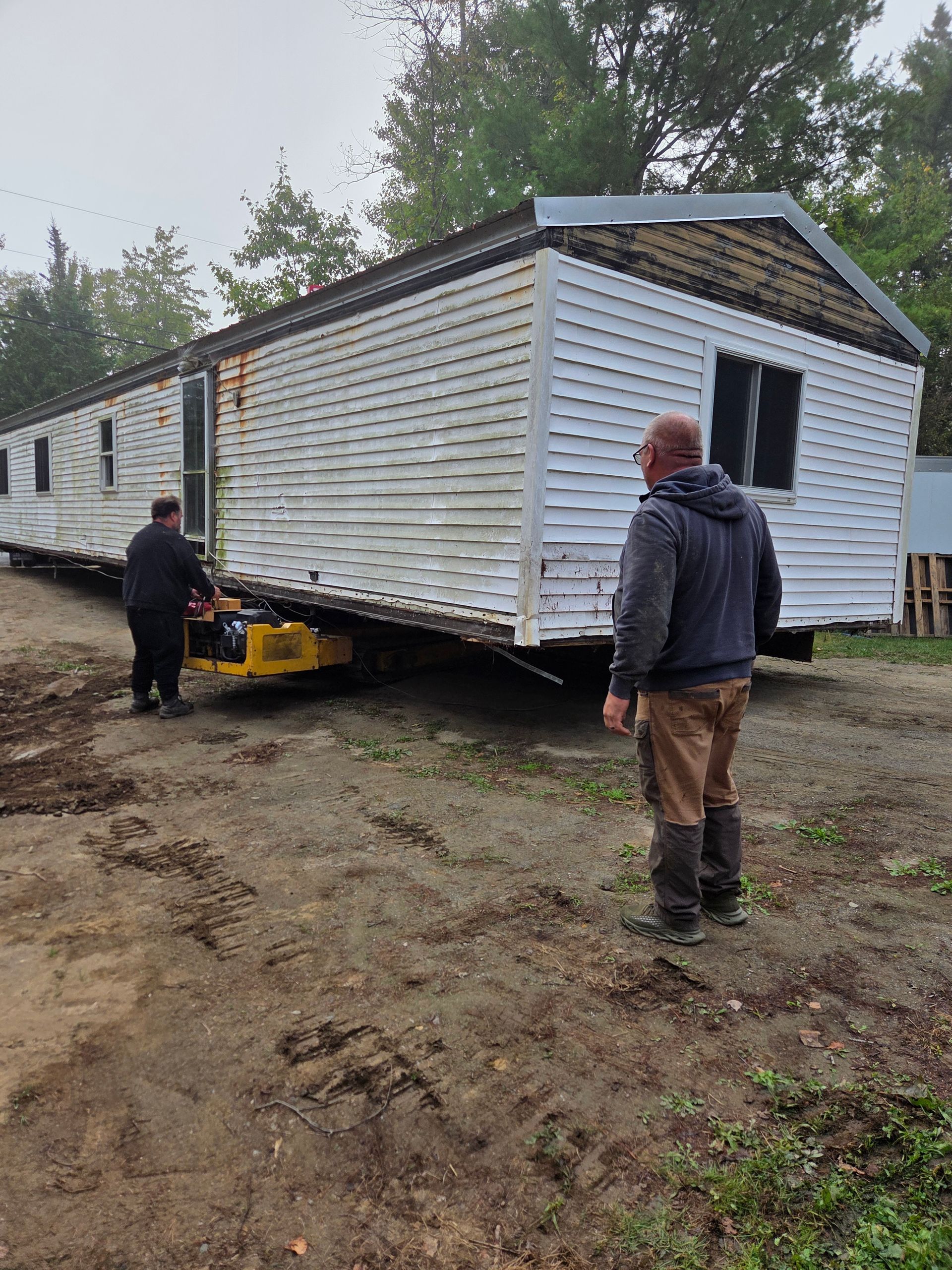 Two people on a gravel lot work to position a white mobile home being moved by a yellow hydraulic trailer.