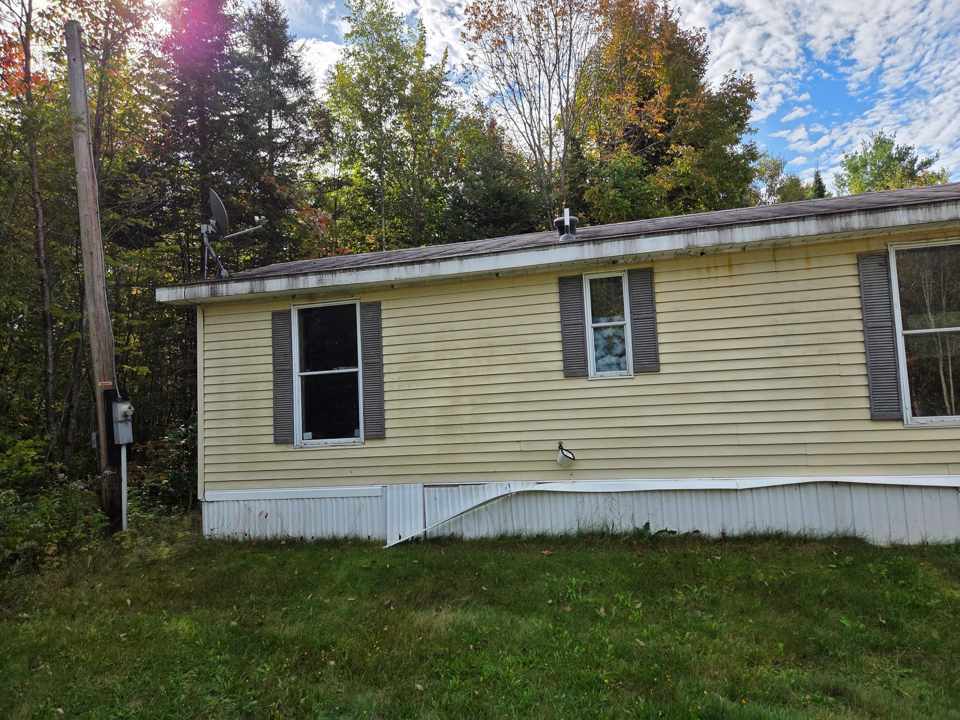 A yellow mobile home with grey shutters and a damaged white skirting panel, set against a backdrop of trees.