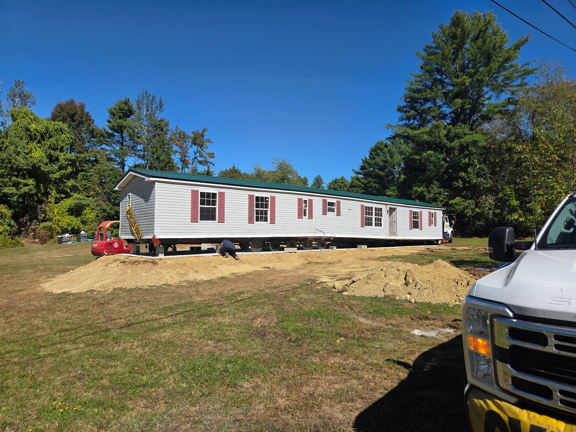 A manufactured home being placed on a gravel foundation in a grassy, tree-lined lot on a sunny day.