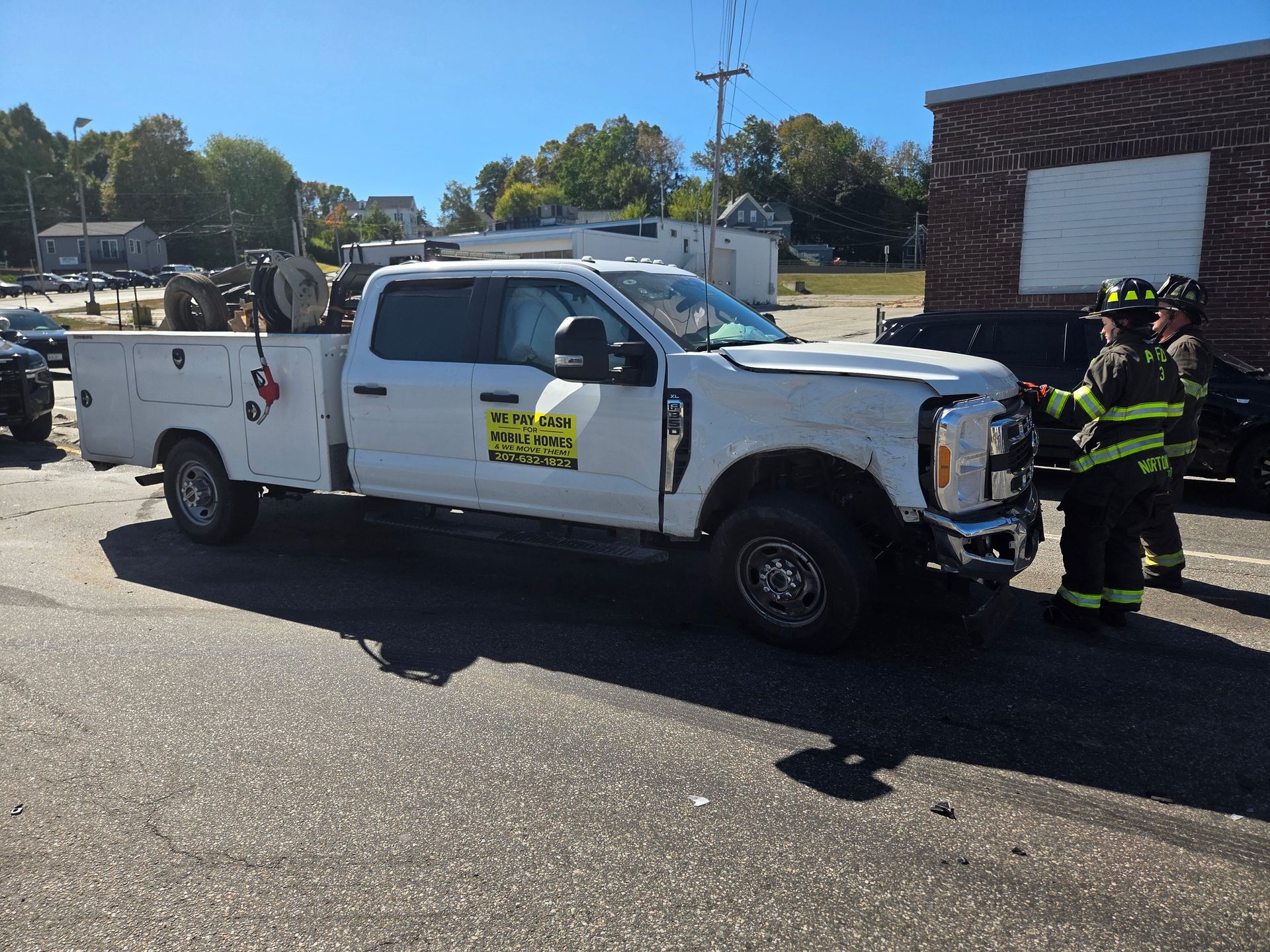 A white service truck with front-end damage parked in an outdoor lot, with two firefighters standing near the bumper.