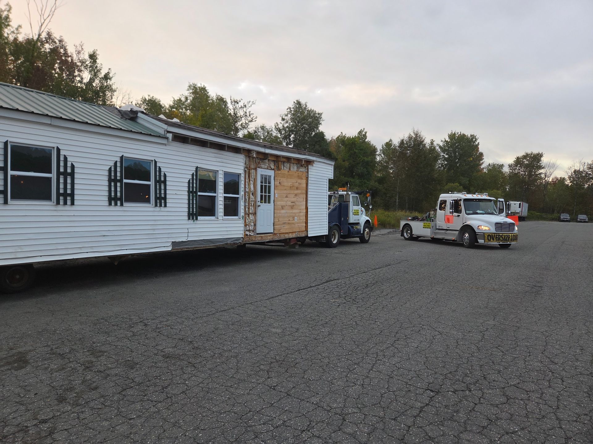 A white mobile home being towed across a gravel parking lot by two trucks under an overcast sky.